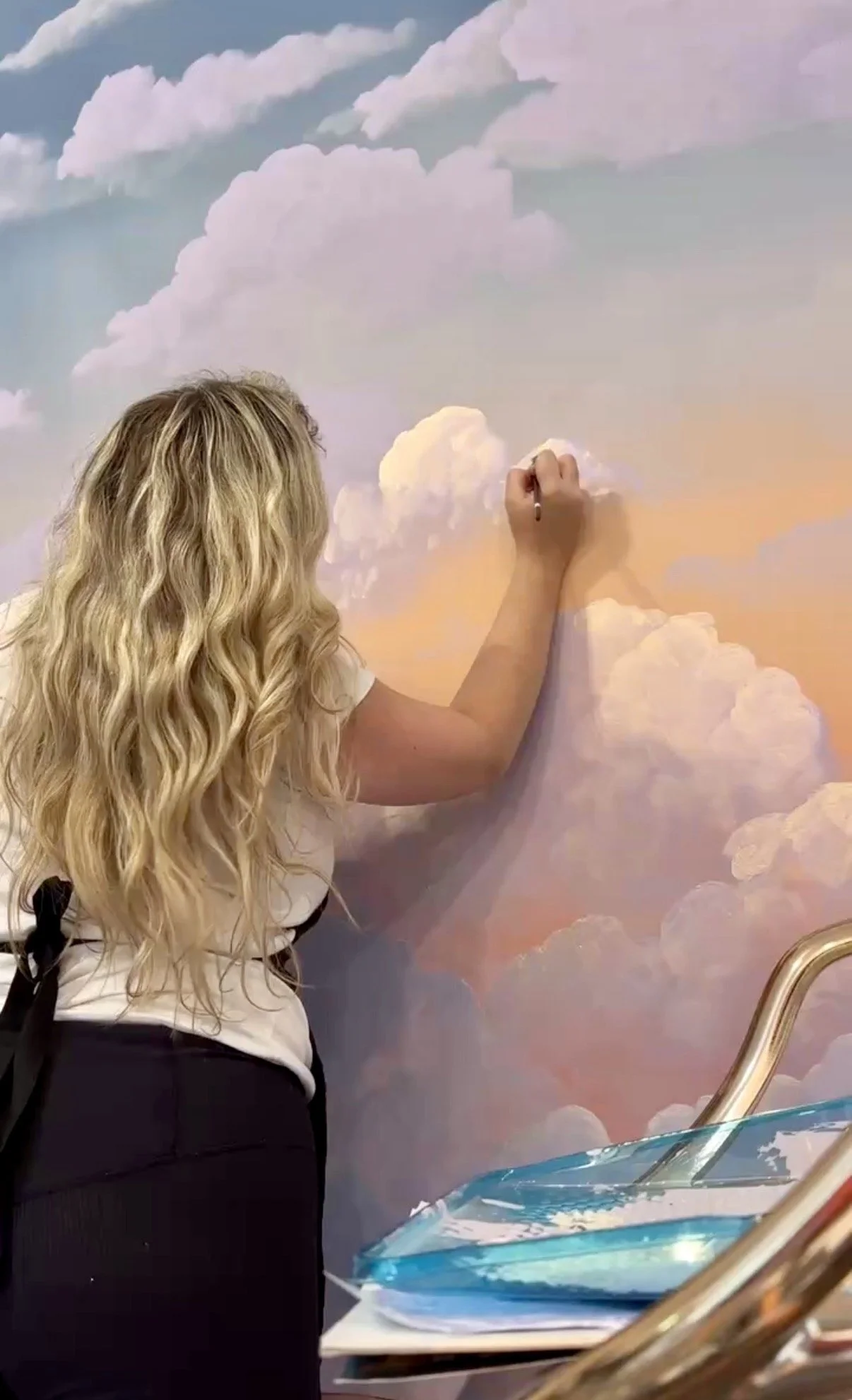 A woman with long, curly blonde hair painting a sky scene on a wall with clouds and a pastel-colored background.