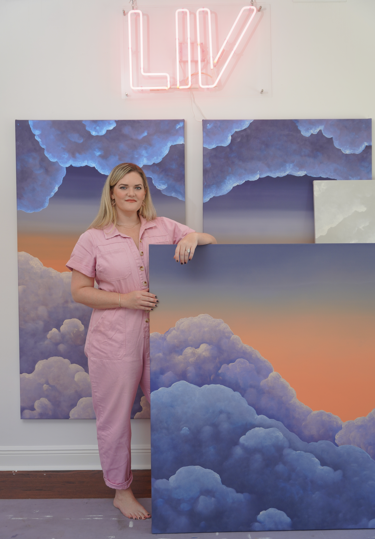 A woman in pink jumpsuit standing barefoot in front of two paintings of clouds and a sunset sky, with a pink neon sign above that says "LOVE."