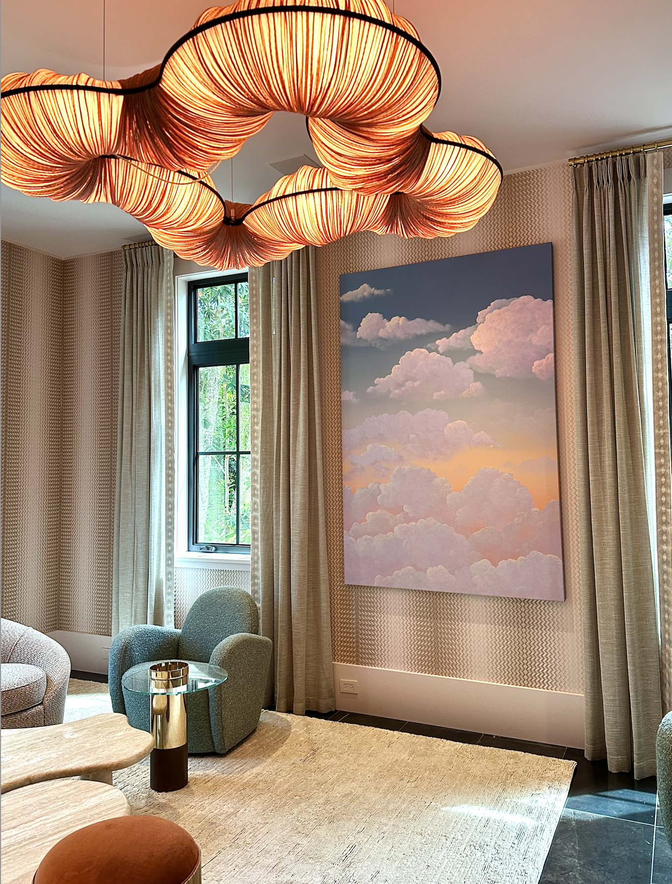 Elegant living room with a cloud-themed wall art, beige curtains, a colorful cloud-shaped ceiling light, and comfortable furniture including a round table, armchairs, and a carpet.