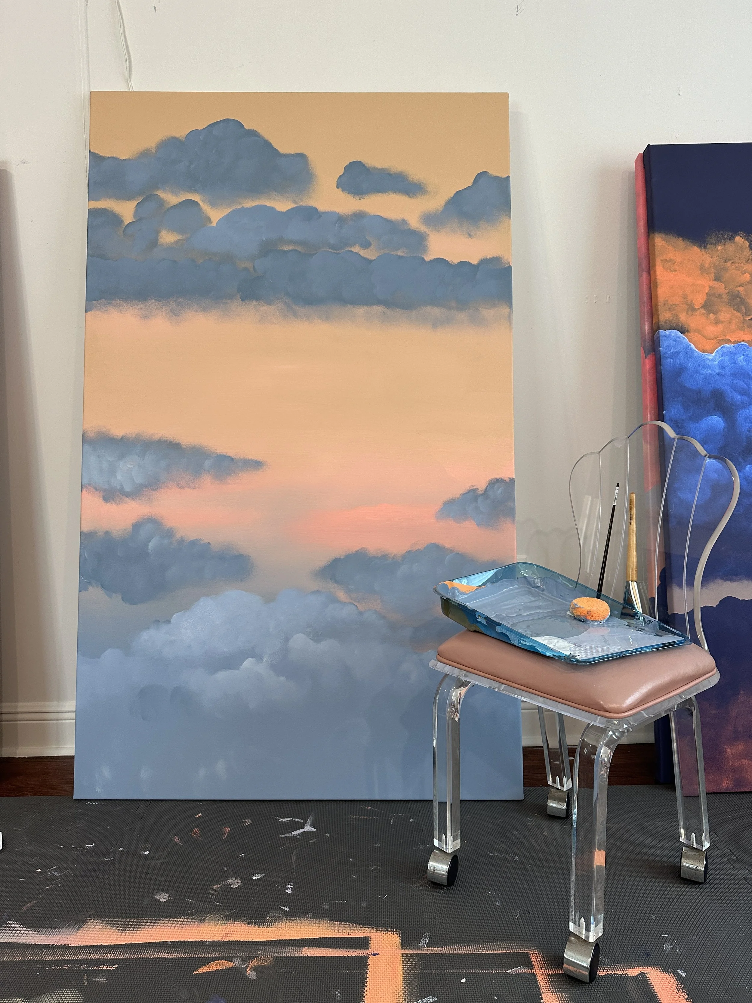 A large canvas with a painted sky scene of clouds at sunset or dawn. The background features blue and gray clouds against a warm orange and pink sky. The canvas is set on a black floor with some paint splatters, and there is a clear acrylic chair wit