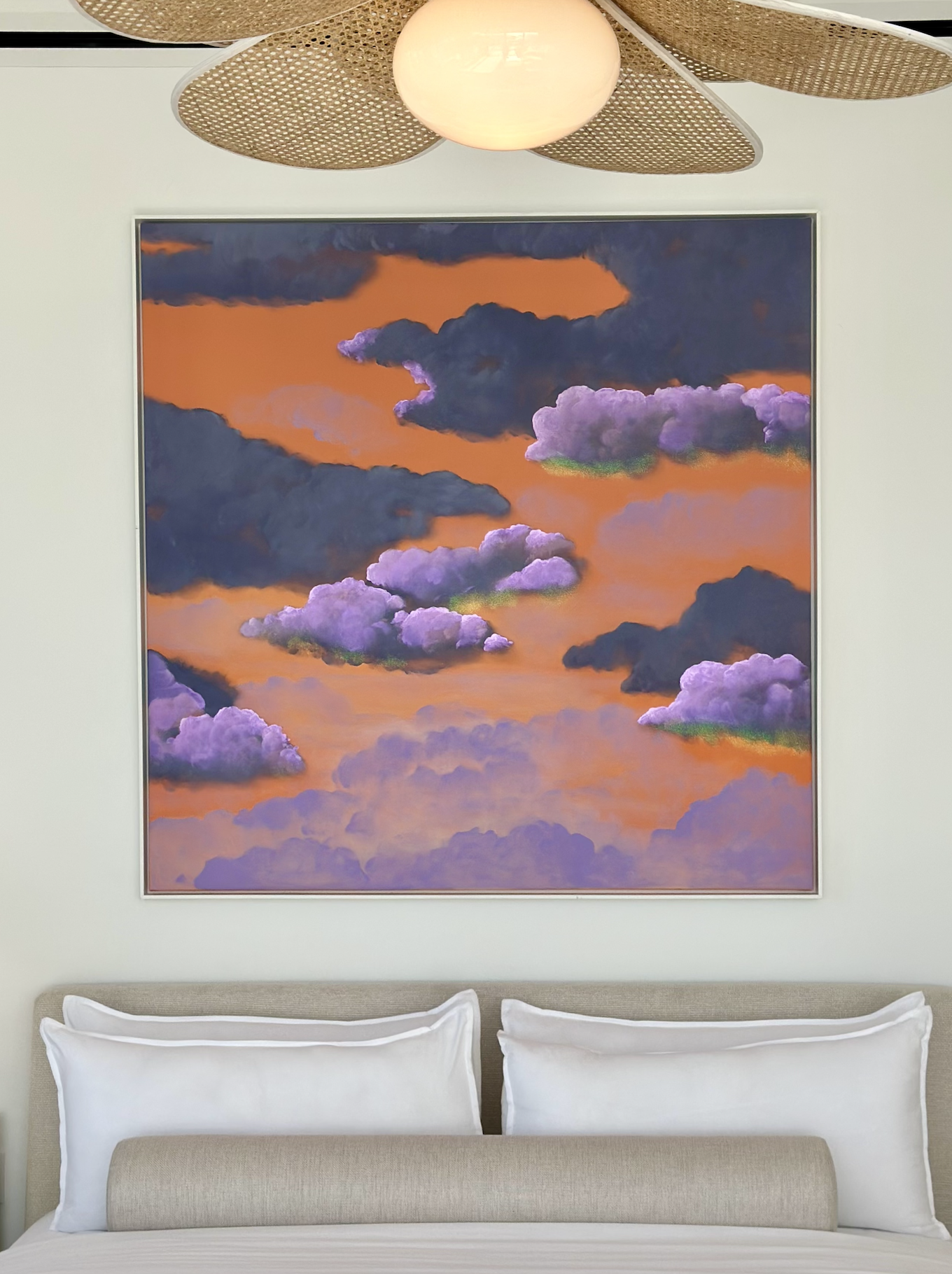 A bedroom with a beige upholstered bed, white pillows, and a colorful painting of clouds in shades of purple, pink, and orange hanging on the wall above the bed. A modern ceiling light fixture is also visible.