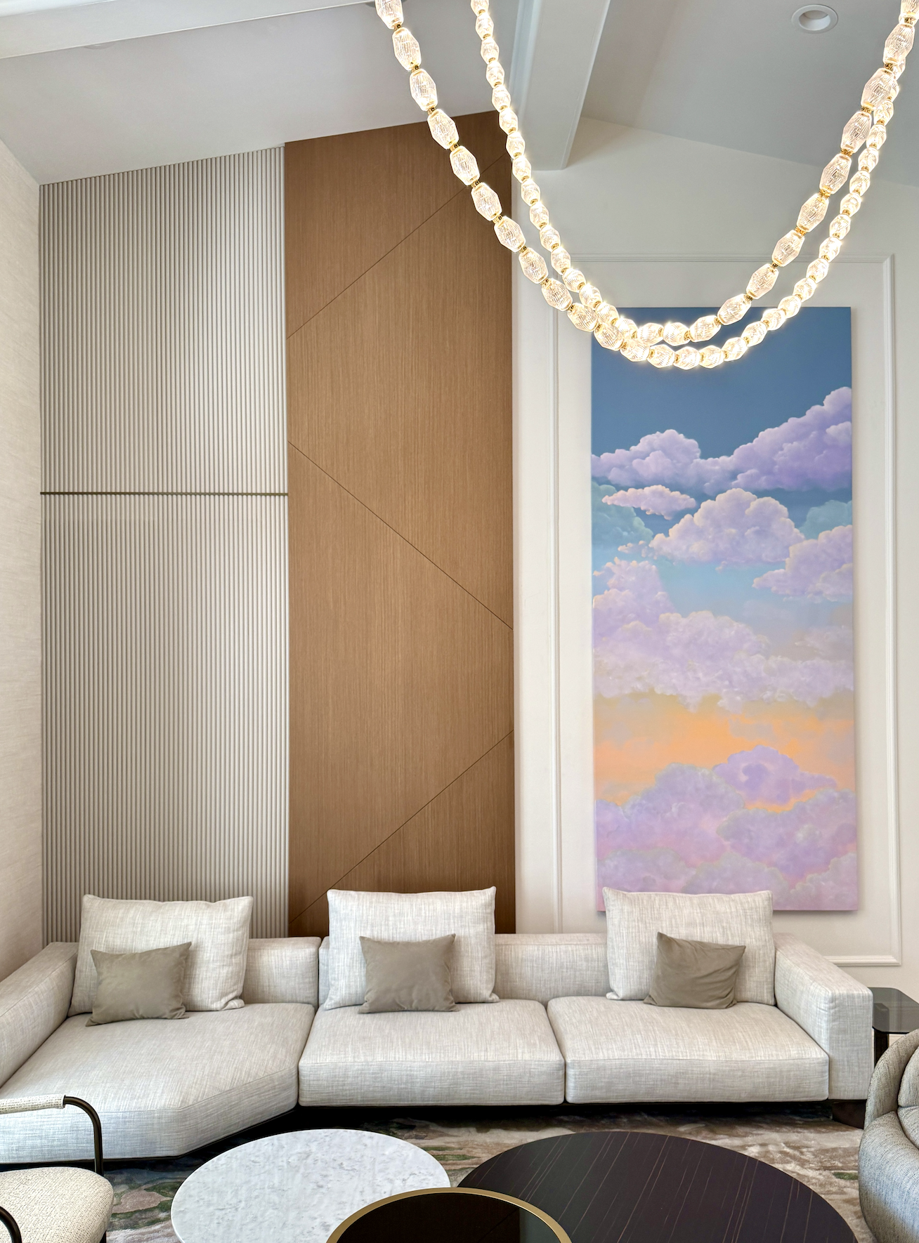 A modern living room with a beige sofa and pillows, a wall with wood and textured panels, a large colorful cloud-themed painting, a chandelier with glass beads, and round coffee tables.