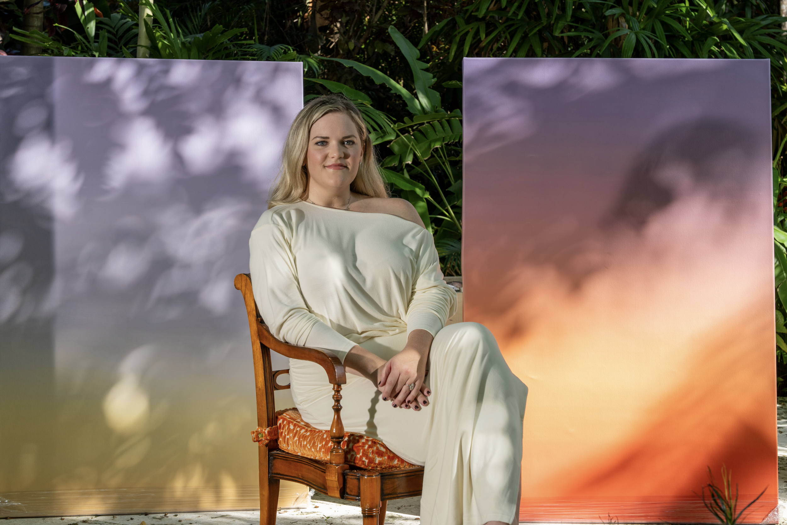 A woman with blonde hair sitting on a wooden chair in front of a backdrop with cloud images. She is wearing a cream-colored off-shoulder top and cream pants, with a slight smile on her face. Green foliage is visible behind the backdrop.