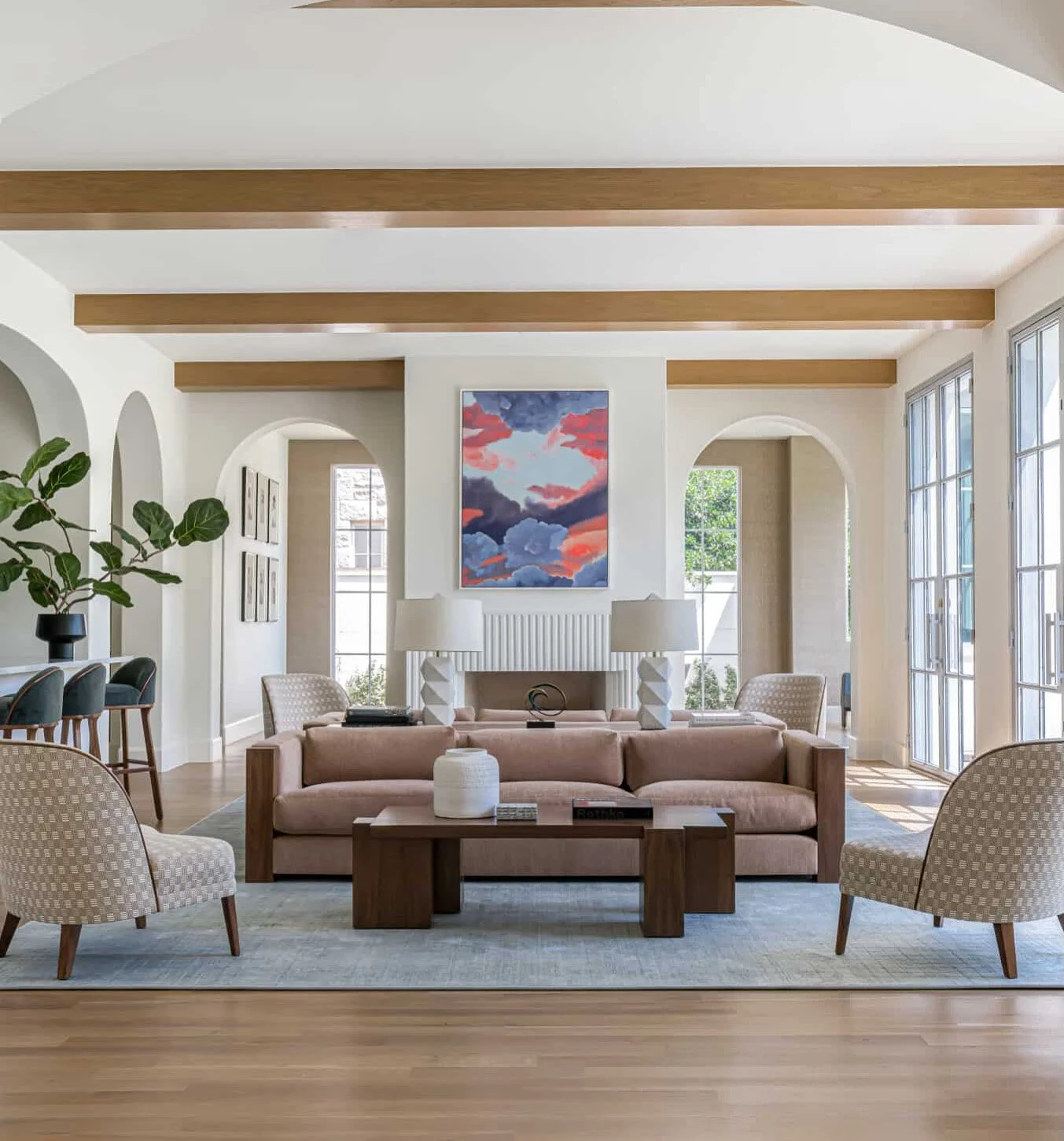 Living room with beige sofa, patterned armchairs, and a wooden coffee table, large windows with natural light, modern paintings on white walls, ceiling beams, and indoor plants.