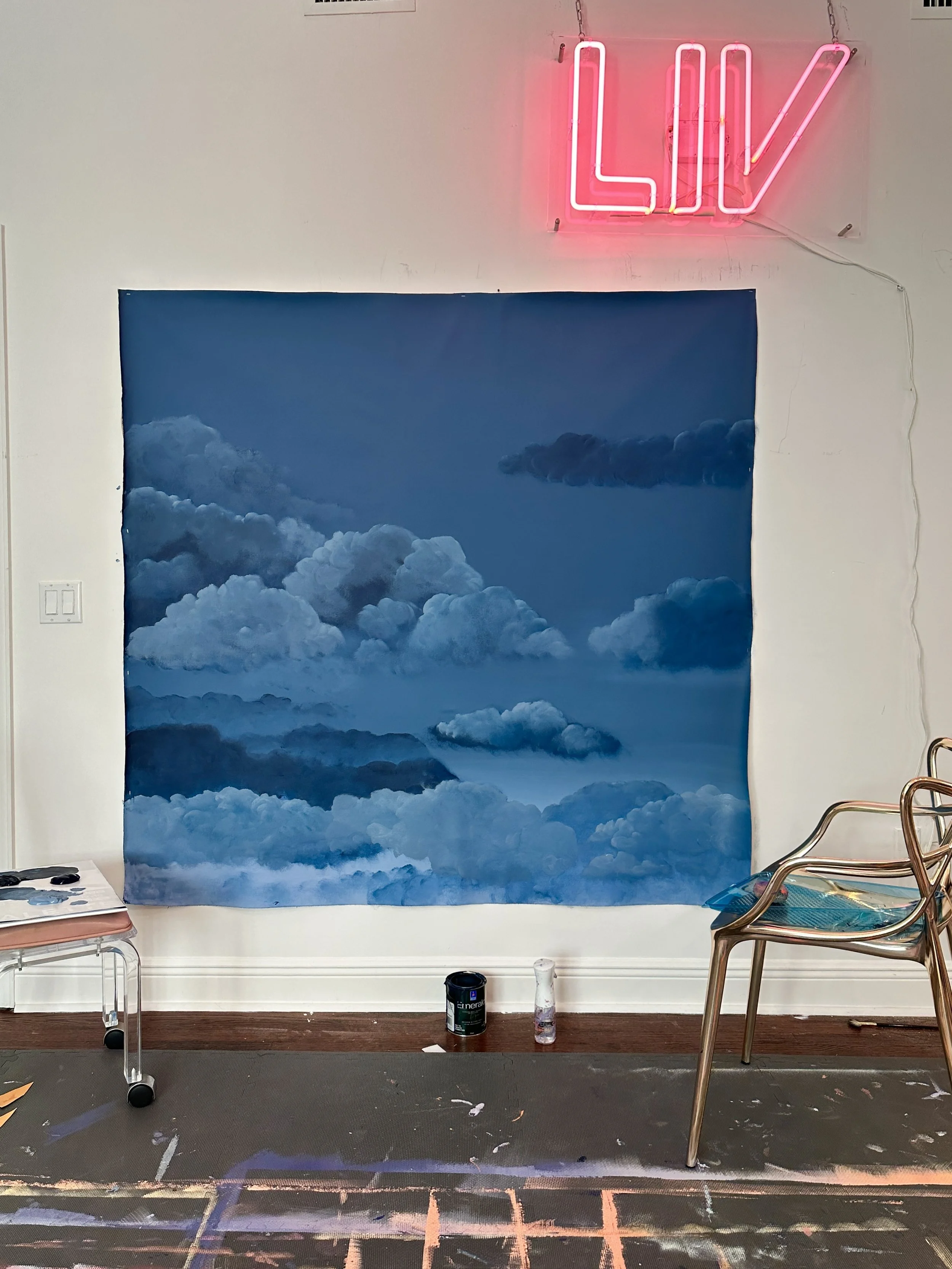 A wall with a large, detailed mural of blue sky with clouds. Above the mural, a pink neon sign spelling 'LOVE' is mounted on the wall. There are chairs, a small table, painting supplies, and a black paint can in front of the wall, indicating an art s