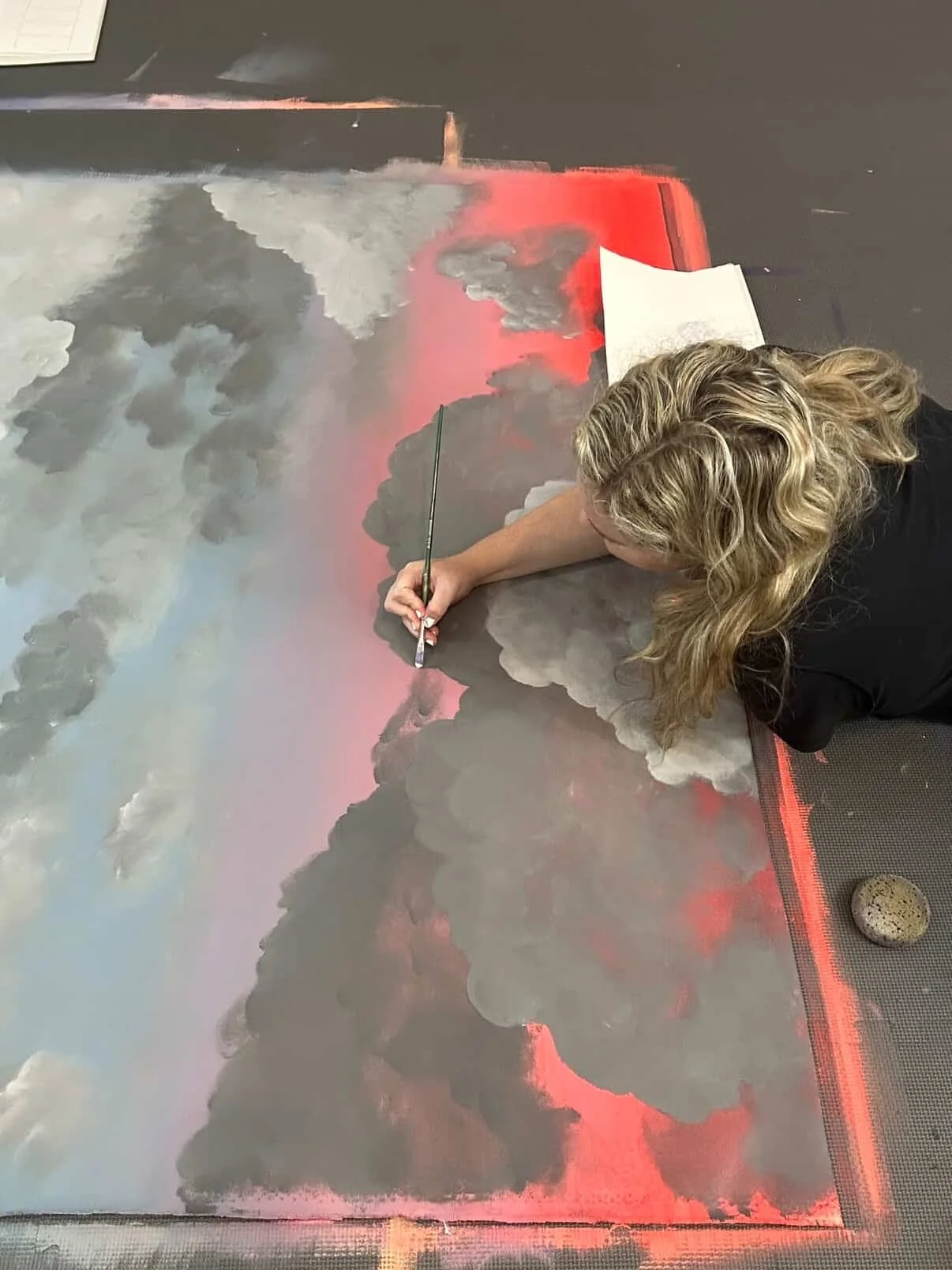Artist creating a cloudscape painting on a large canvas, using shades of gray, white, black, and red, in an indoor setting.