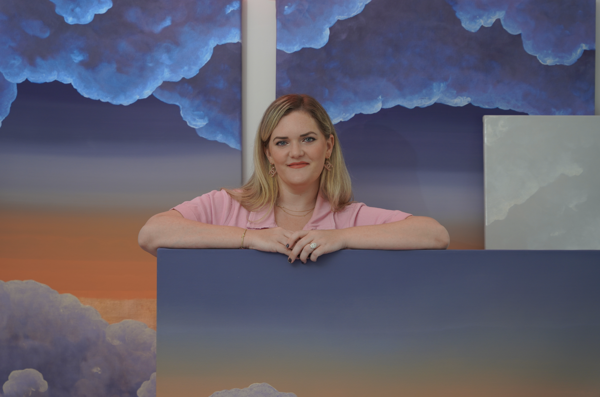 A woman with blonde hair and blue eyes leaning on a blue counter with her arms crossed. She is wearing a pink shirt, earrings, and rings. Behind her is a painted backdrop of blue clouds and a sky with shades of orange and purple.