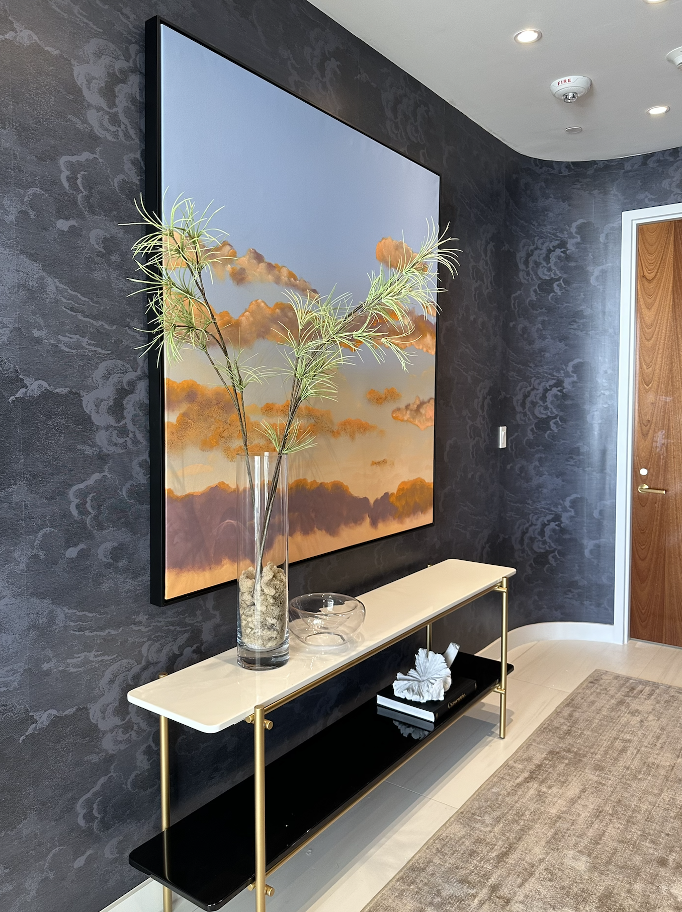 Interior view of a modern living space with a black wall featuring a painting of orange-clouded sky, a beige and black console table with gold legs, a glass vase with decorative branches, a bowl, and stacked books with a decorative object.