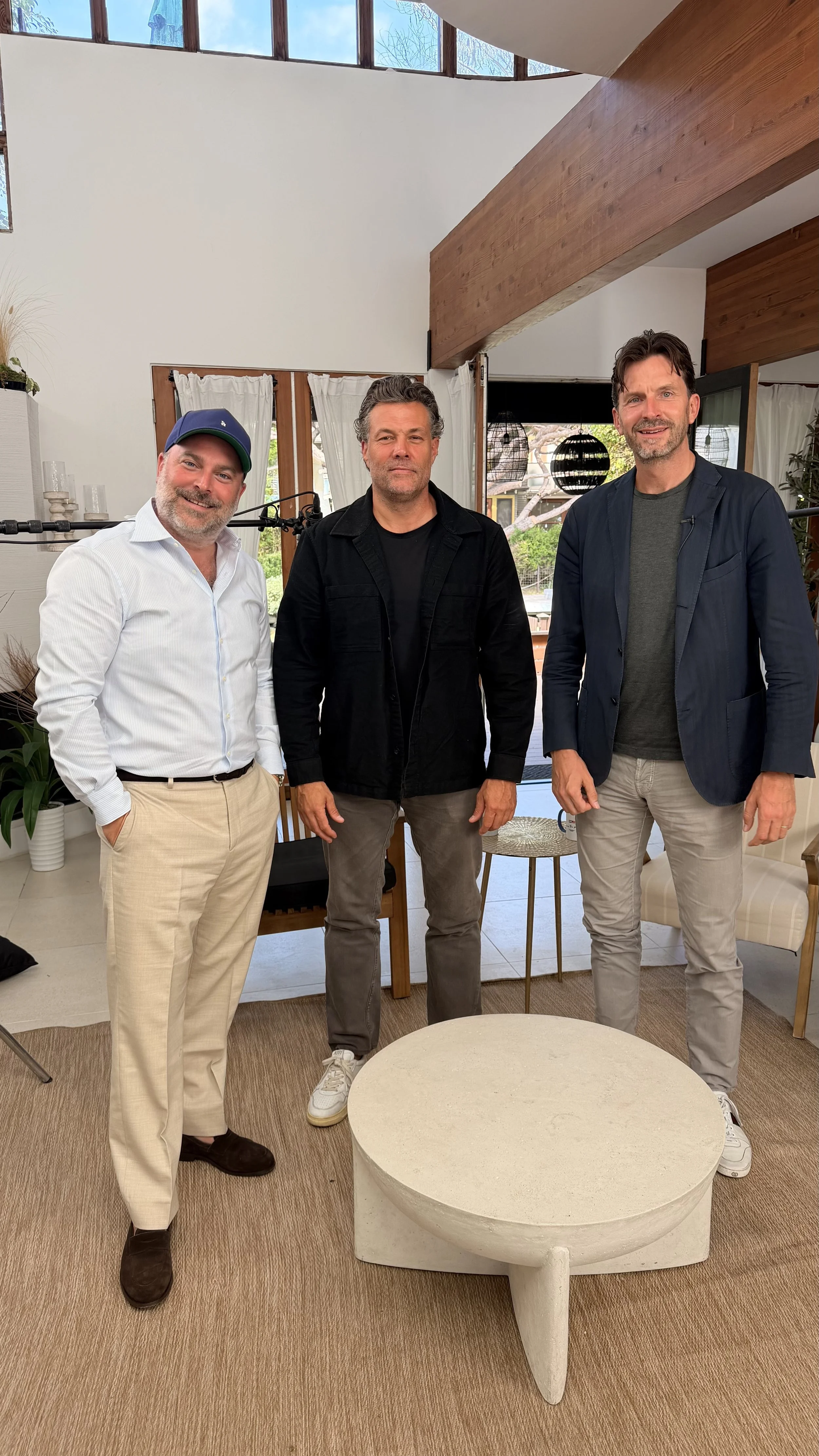 Three men standing inside a modern house, posing for a photo. The man on the left is wearing a light-colored shirt, beige pants, and a cap. The man in the middle is wearing a black jacket and gray pants. The man on the right is wearing a dark blazer over a gray shirt and light-colored pants. There is a round, white table in the foreground, and the background features large windows, curtains, and decorative lighting fixtures.