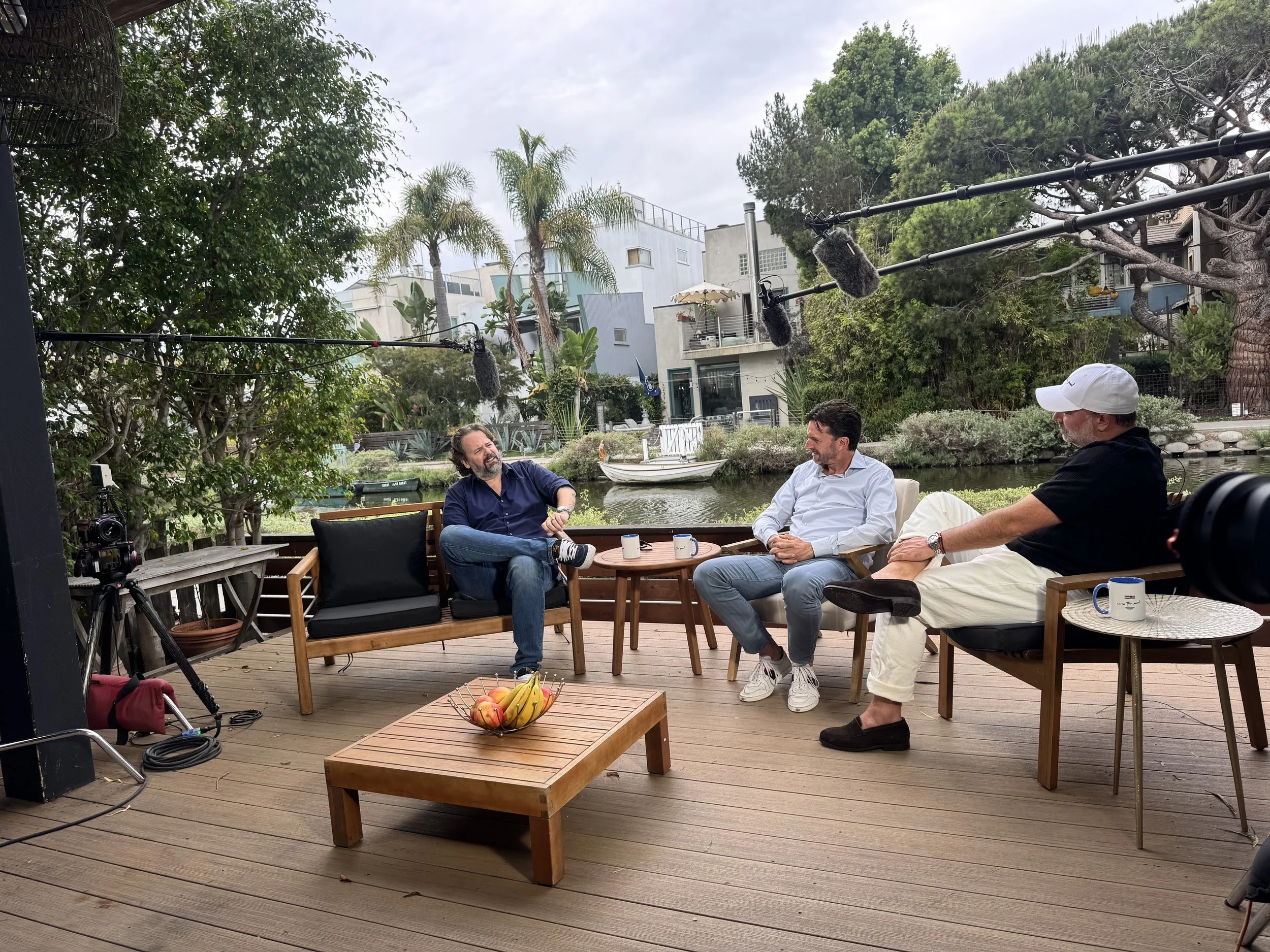 Three men sitting outdoors on a deck, engaged in conversation, behind a river with boats, trees, and modern houses, with recording equipment overhead.