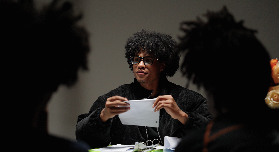 Young man with curly hair and glasses sitting at a table, holding a paper, with two people in the foreground.
