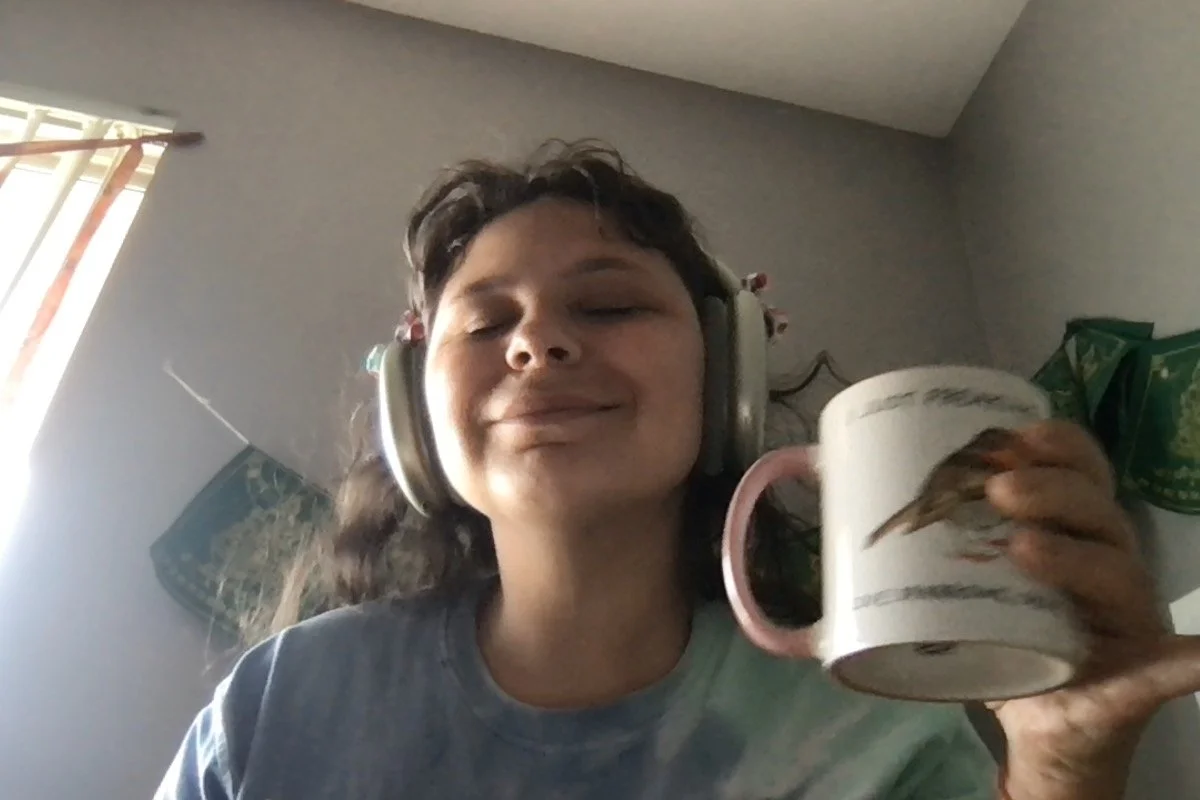 Young woman with curly hair wearing headphones, holding a mug with bird design, smiling with closed eyes in a room with a window covered partly by blinds. And she's so pretty!