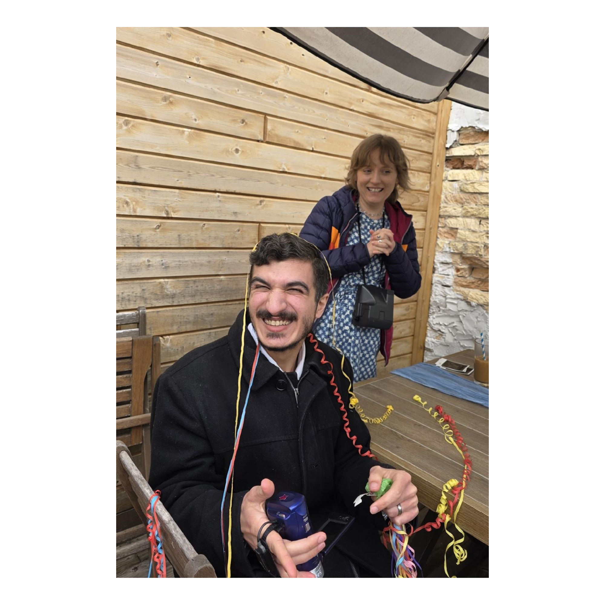 Group of people celebrating with colorful streamers, holding party poppers, and smiling, with a wood-paneled wall and stone wall in the background.
