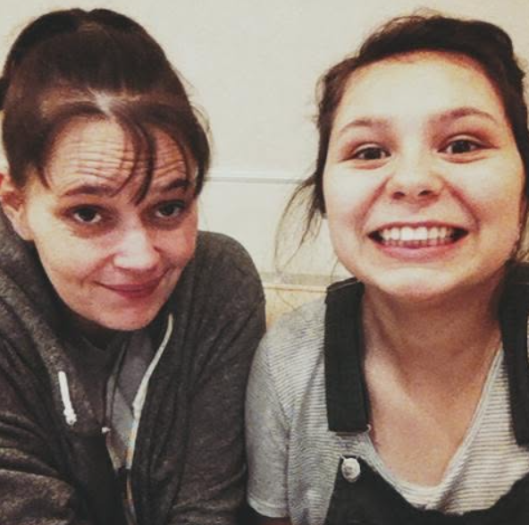 Two women taking a selfie, smiling at the camera. One wears a gray shirt with overalls, and the other has a gray hoodie with white earphones.