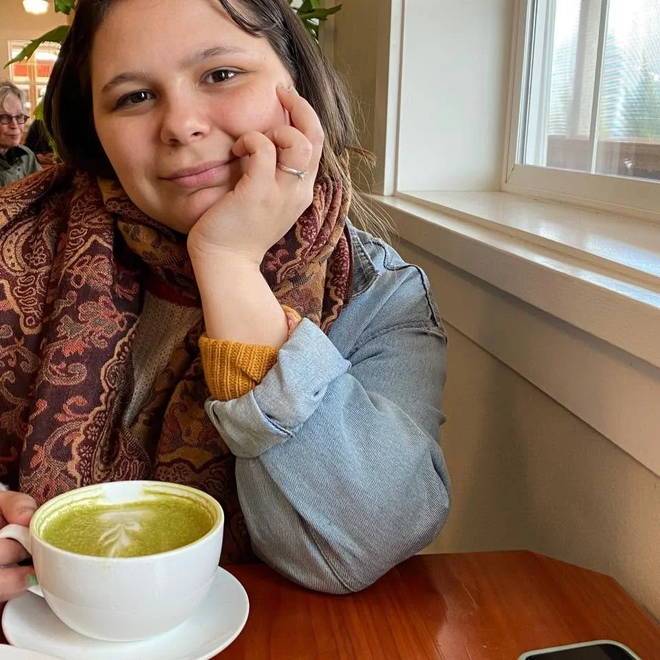 A young woman with brown hair resting her face on her hand, sitting at a table near a window with a cup of matcha latte in front of her. She is wearing a patterned scarf, a yellow sweater, and a gray jacket.