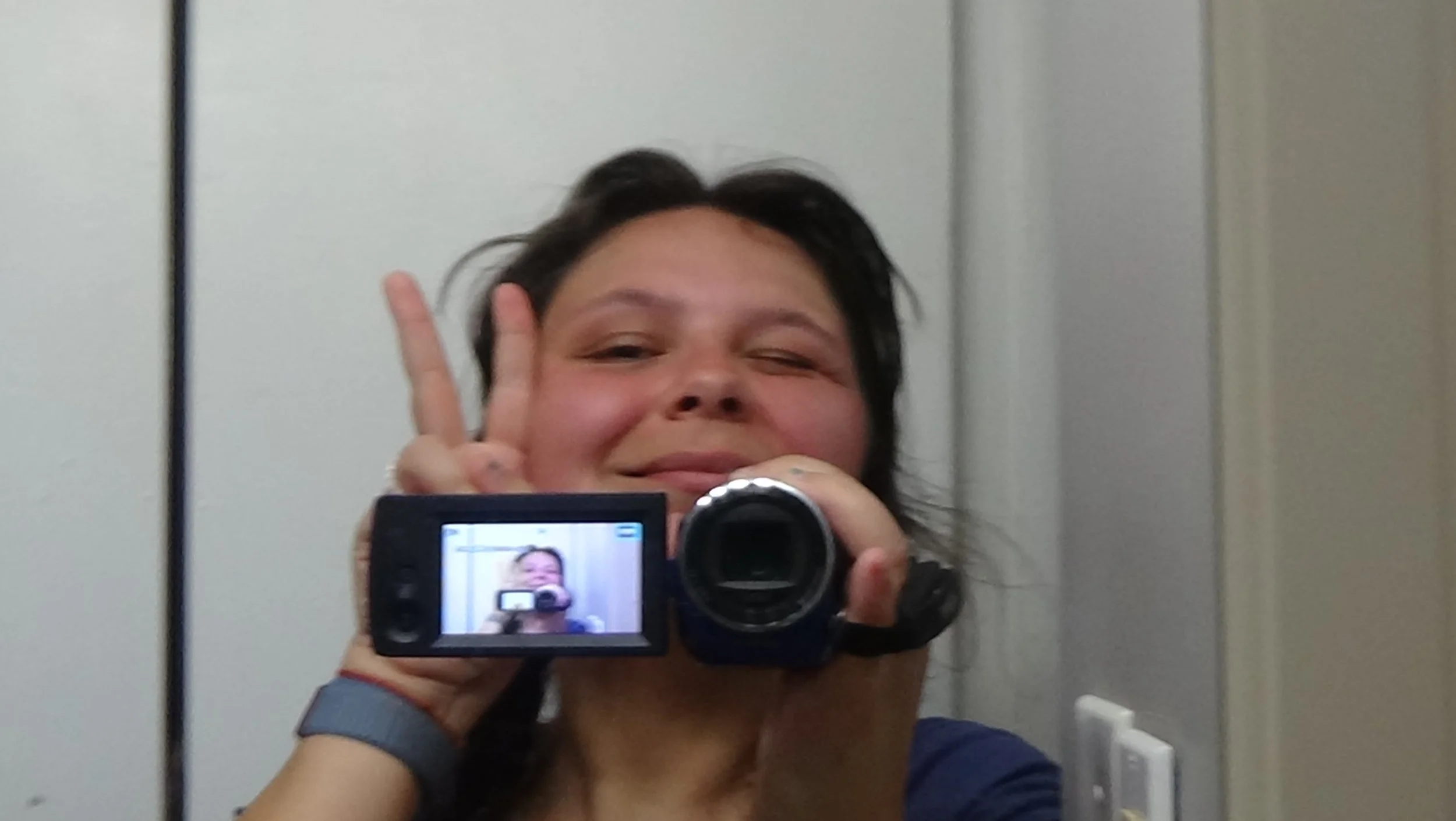 Person taking a selfie with a camcorder in front of a mirror, showing their face and making a peace sign with one hand.