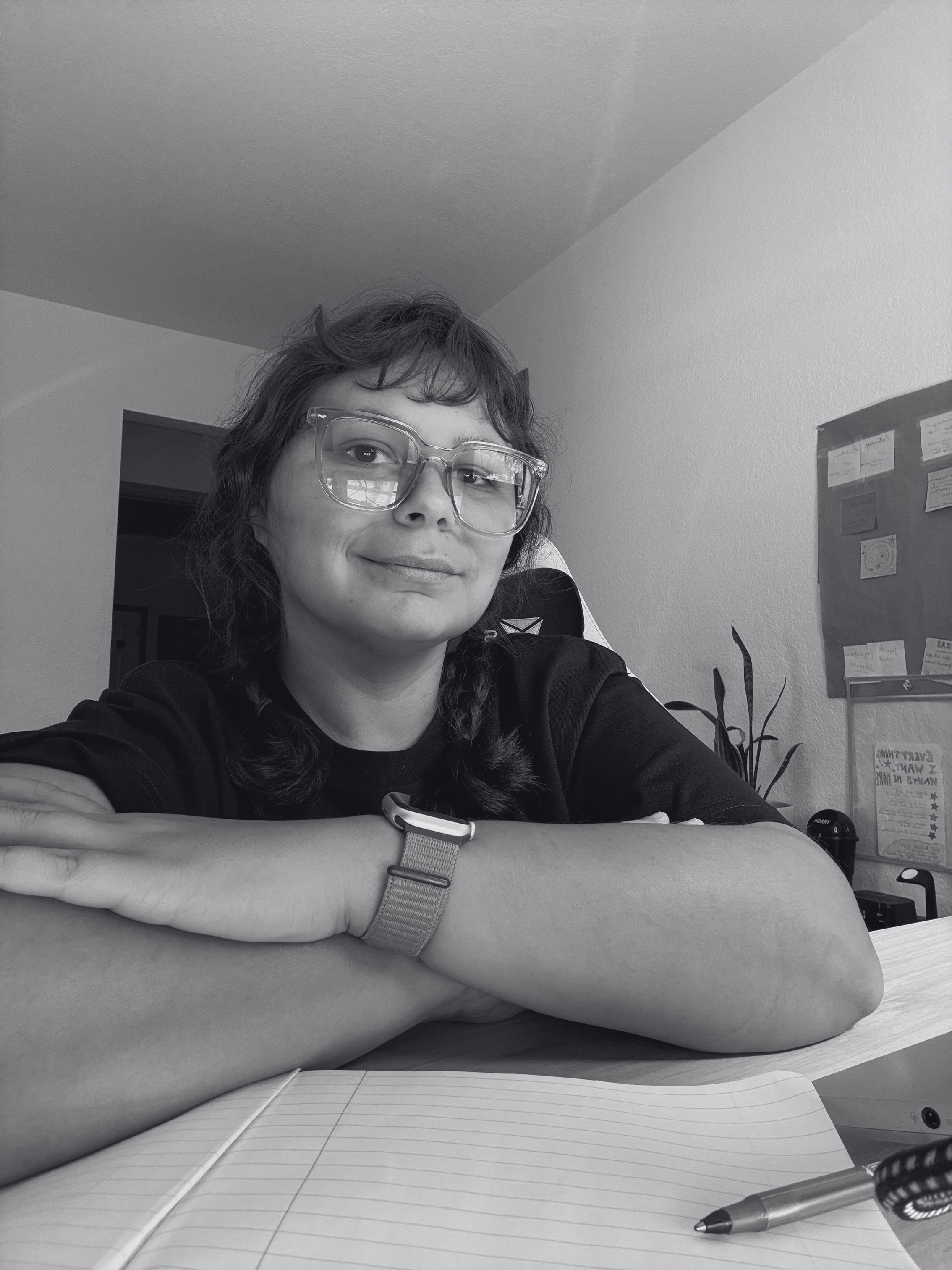 A person with wavy hair and glasses smiling while resting their arms on a table, with a notebook and pen in the foreground. In the background, there are notes or posters on the wall.