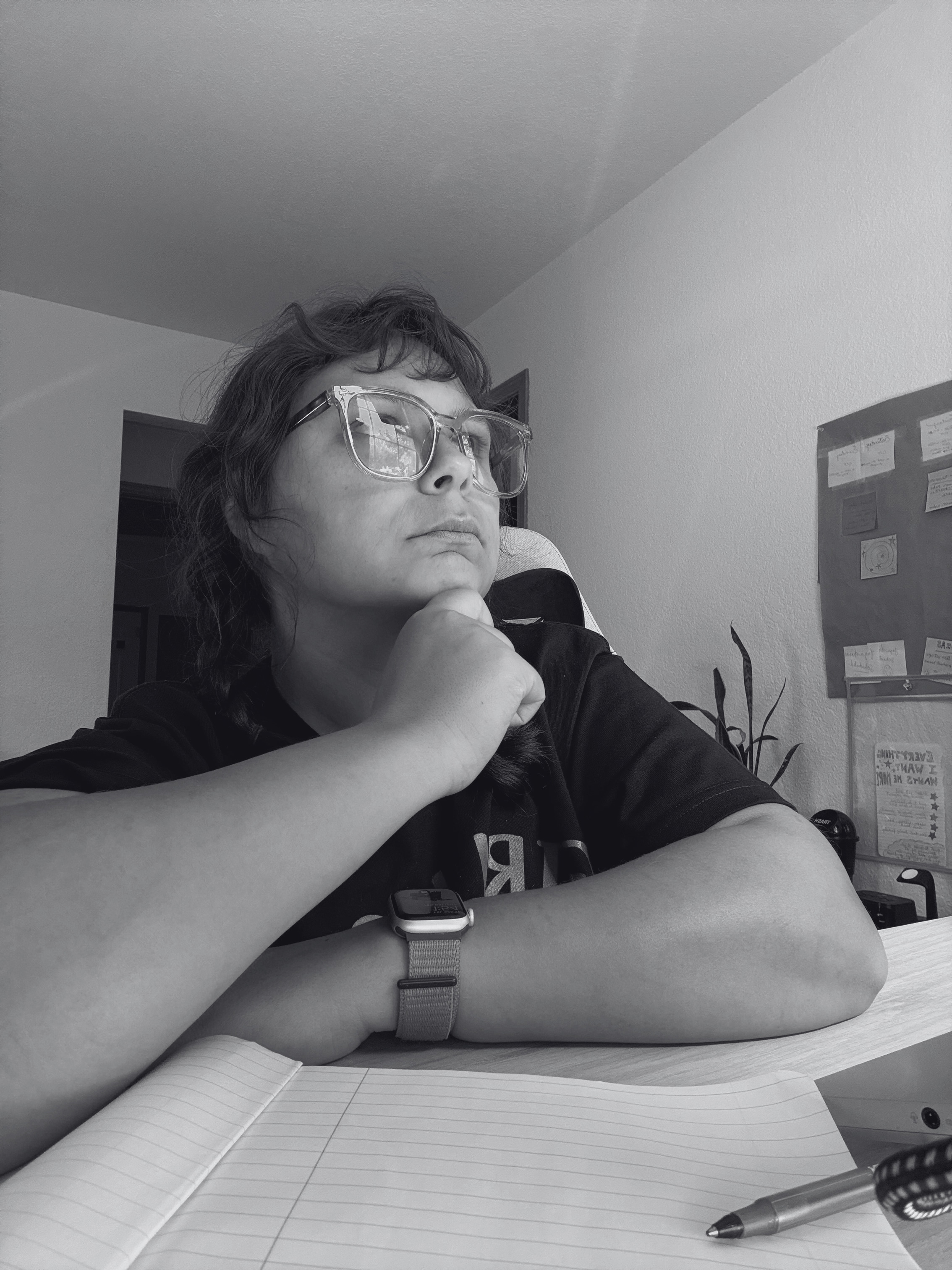 A person wearing glasses and a watch, sitting at a desk with an open notebook and a pen, resting their chin on their hand, with a bulletin board on the wall in the background.