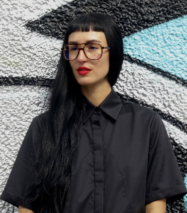 Woman with straight black hair, glasses, and red lipstick standing in front of a textured wall with black, white, and blue geometric shapes.