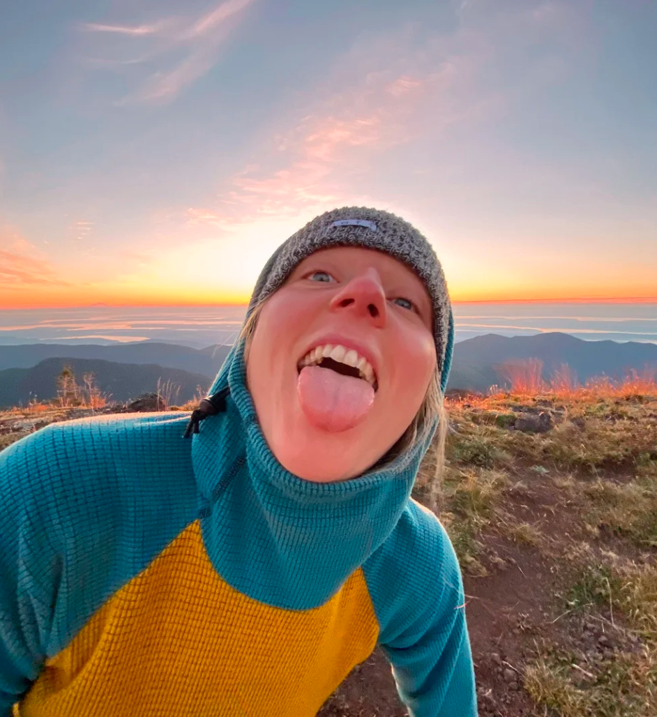 A person in a teal and yellow jacket and gray beanie making a silly face with their tongue out, outdoors on a mountain at sunset with a colorful sky and distant hills in the background.