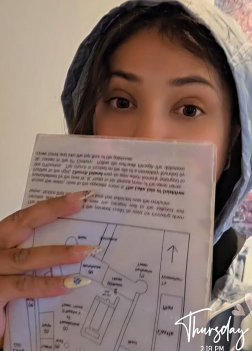 Young woman wearing a hooded jacket holding a laminated map or diagram in front of her face, with only her eyes and forehead visible, indoors with a light-colored wall behind her.