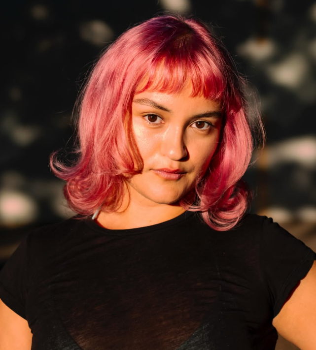 Young woman with pink, shoulder-length hair and bangs, wearing a black top, standing outdoors during sunset.