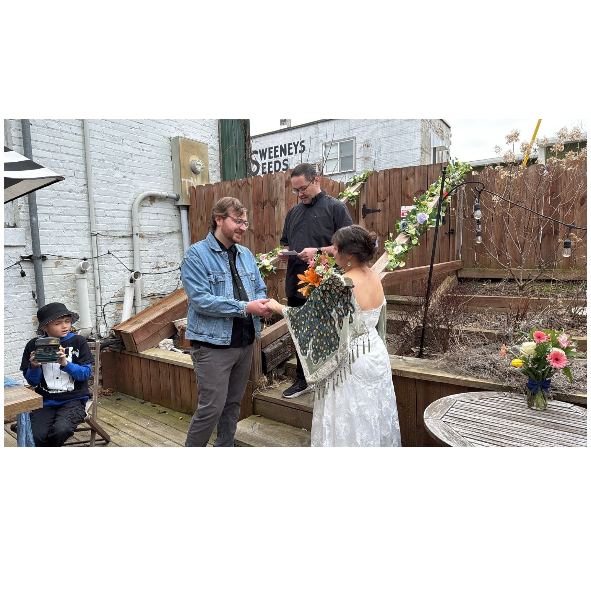 An outdoor wedding ceremony with a bride and groom holding hands, a officiant, and a few guests present. The setting includes a wooden fence, string lights, and floral decorations.