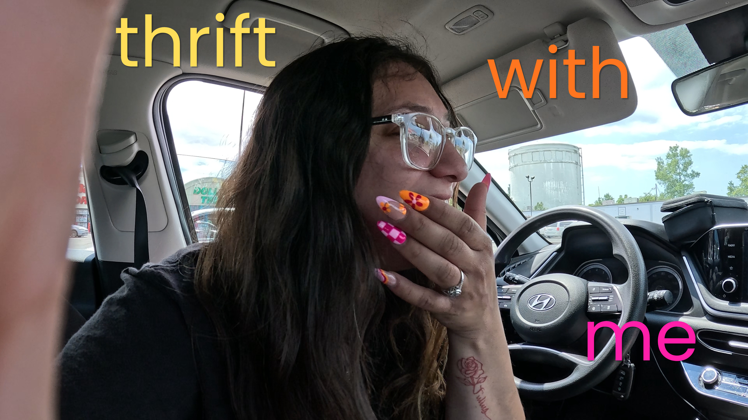A woman with long dark hair and colorful nail polish is sitting in the driver's seat of a car, touching her face with her hand. She is wearing glasses and has tattoos on her arm. The background shows a parking lot with a large white water tower and some trees. Overlaid text reads "thrift with me."