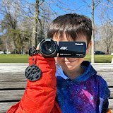 Child in a red jacket holding a small black camera, taking a photo in an outdoor park.