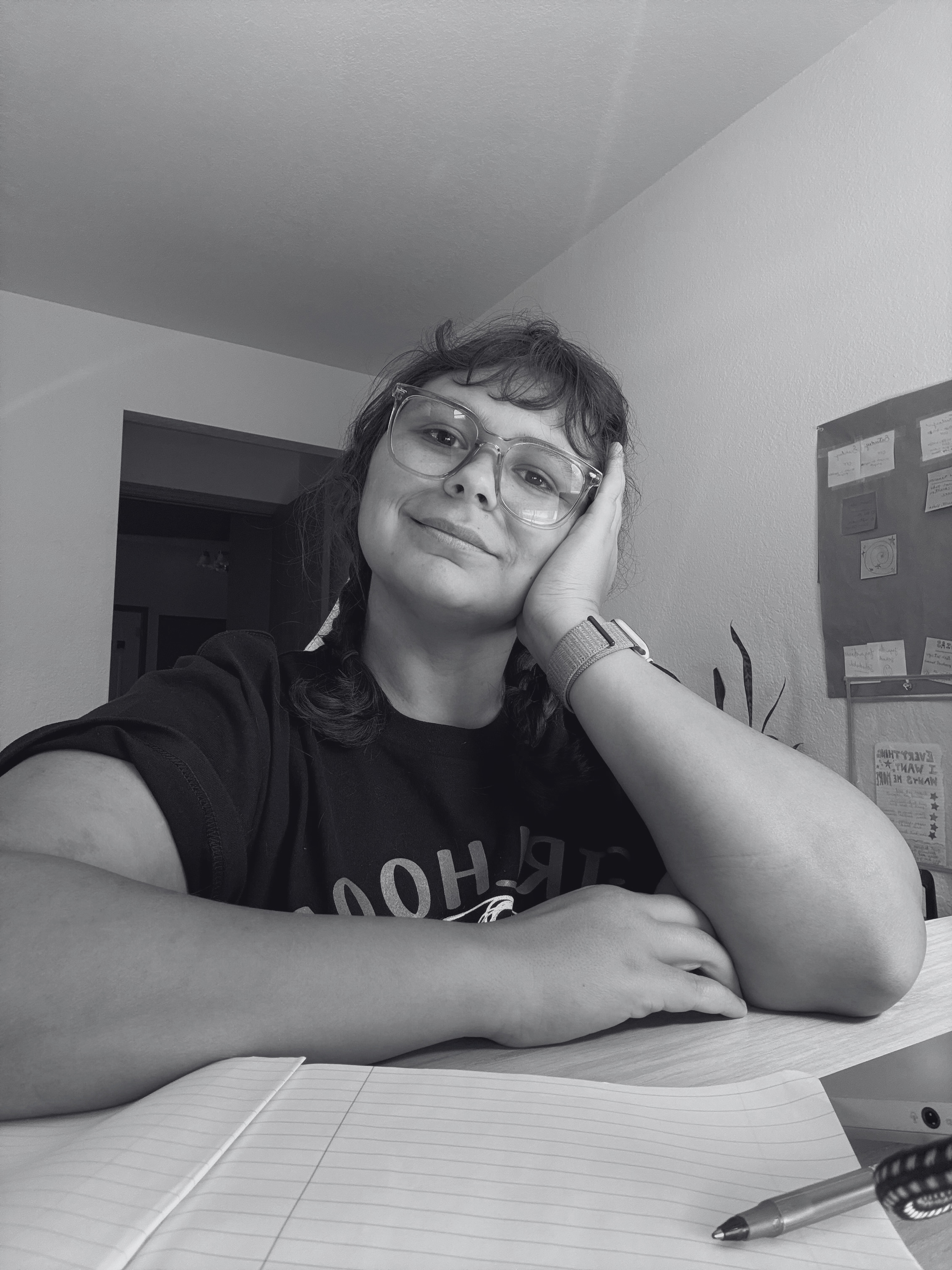 A woman with glasses and wavy hair sitting at a table, resting her head on her hand, smiling at the camera, with a notebook and pen in the foreground, in a room with wall decorations.