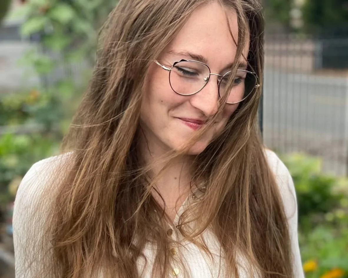 A young woman with long, wavy brown hair and glasses smiling outdoors, in front of a garden with greenery and flowers.