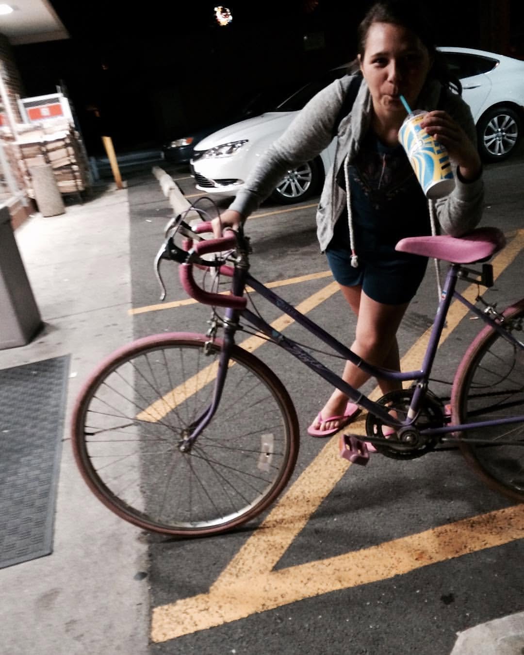 A woman in casual clothing and flip-flops is holding a pink bicycle while drinking from a striped cup with a straw in a parking lot at night.