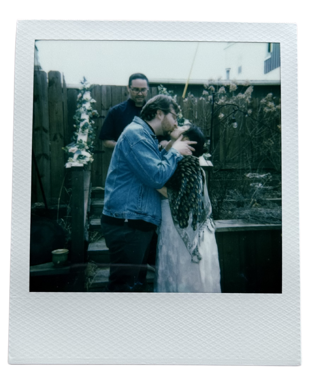 A Polaroid photo of three people outdoors in a backyard with a wooden fence, plants, and a garden bed. One person with glasses wearing a denim jacket is kissing a woman, while another man with glasses and a beard stands in the background.