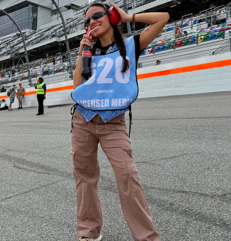 Smiling woman at a race track wearing sunglasses, a racing bib with the number 20, holding a soda bottle and headphones, with a grandstand in the background.
