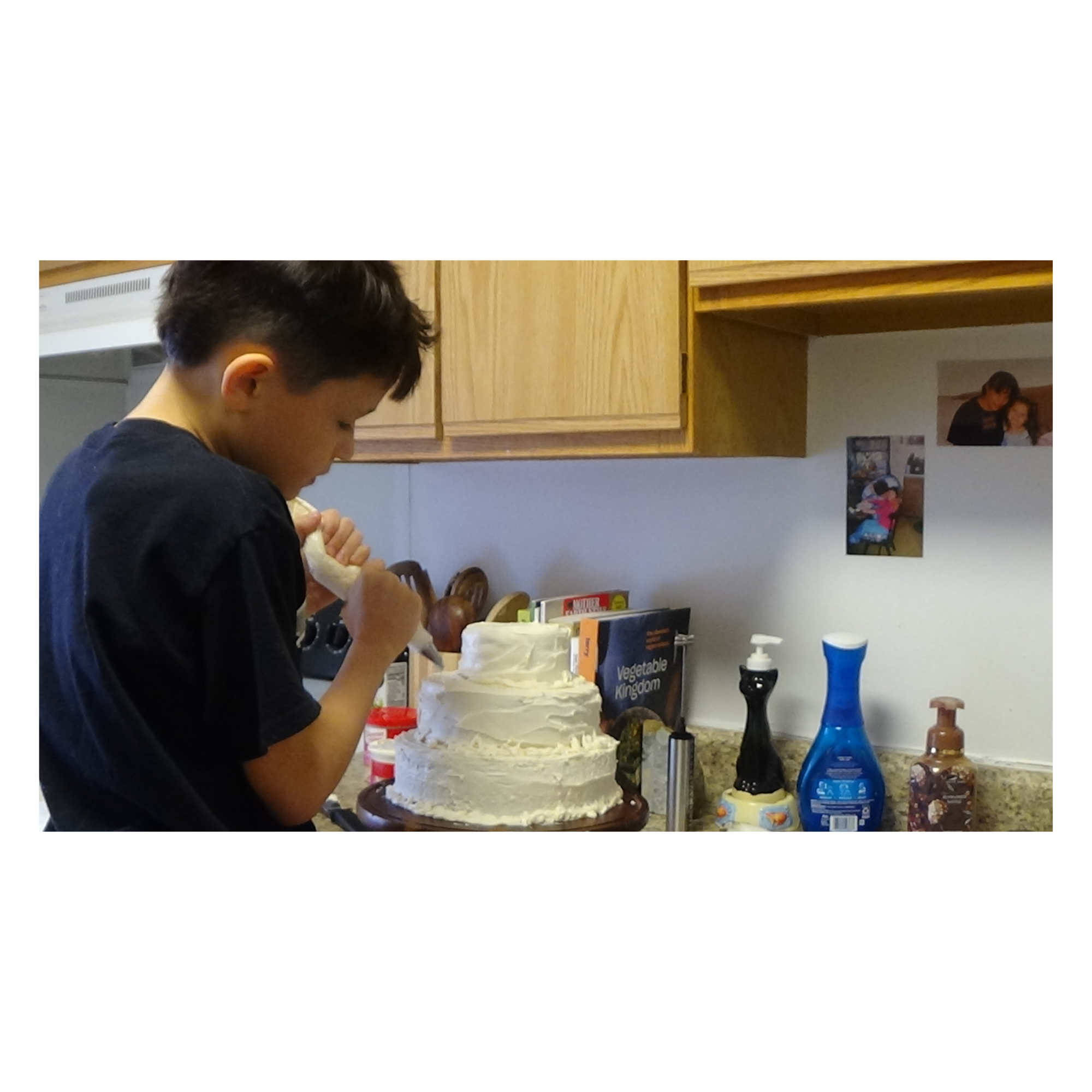 A young man in a black shirt is in a kitchen decorating a large layered cake with white frosting. The kitchen has wooden cabinets and various baking supplies on the counter, including a cake stand, a book titled 'Vegetable Kingdom,' and several bottles and utensils.