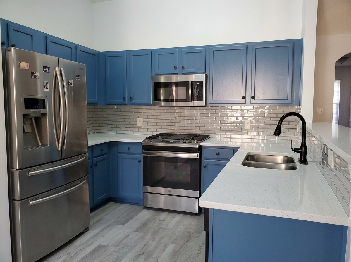 Kitchen with blue cabinets, stainless steel refrigerator, microwave, oven, and a white countertop with a built-in sink and black faucet.