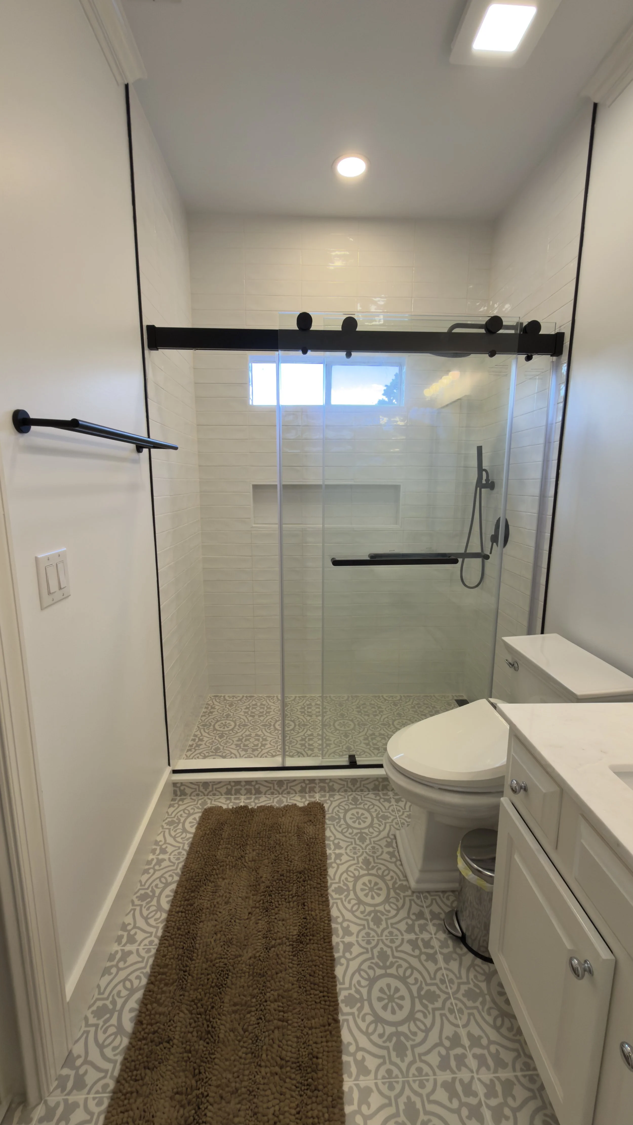 Bathroom with patterned floor tiles, brown bath mat, glass shower with black hardware, white vanity, and a toilet.