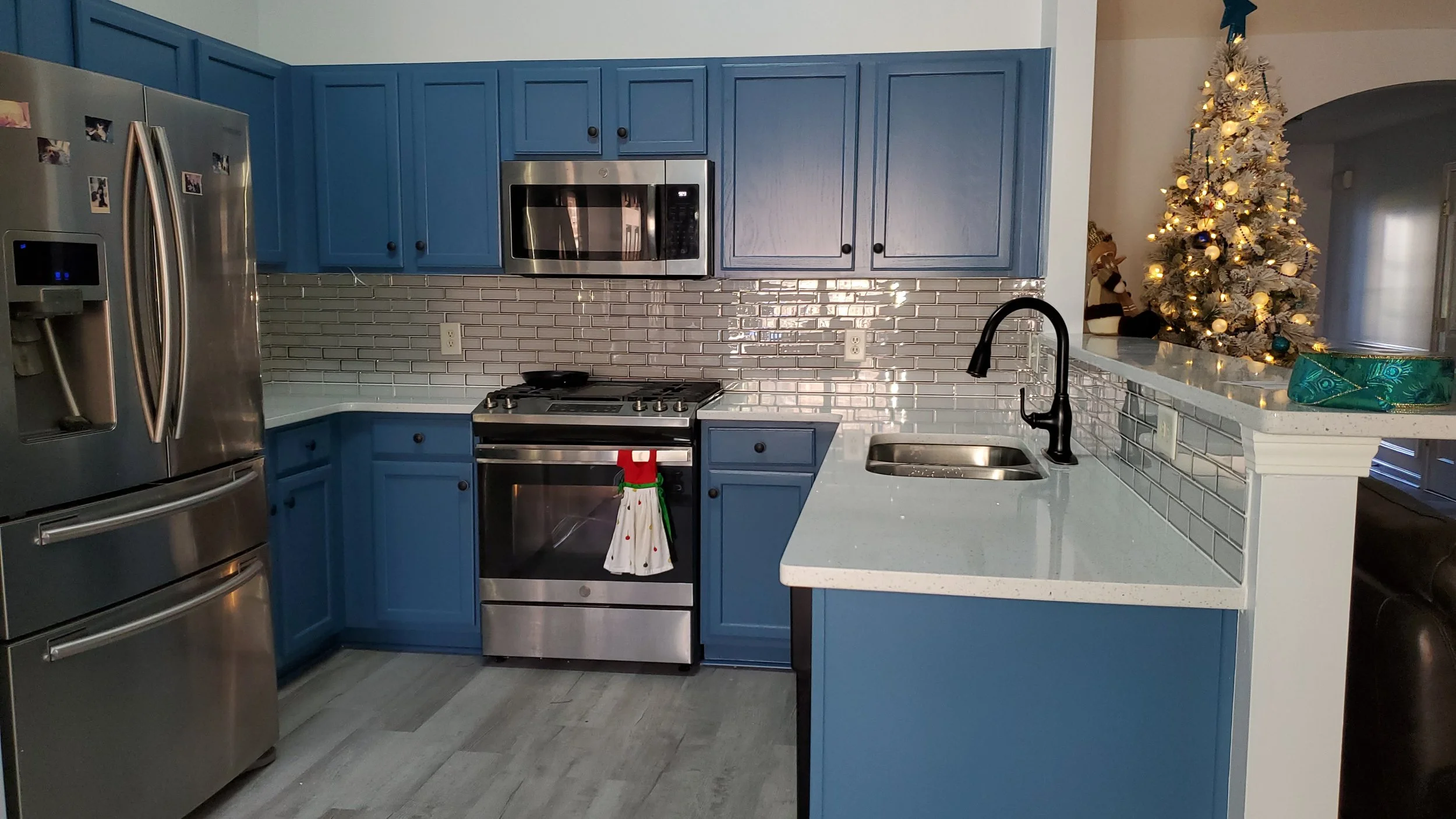 Kitchen with blue cabinets, stainless steel appliances, a backsplash with gray tiles, and a Christmas tree with ornaments and lights in the background.