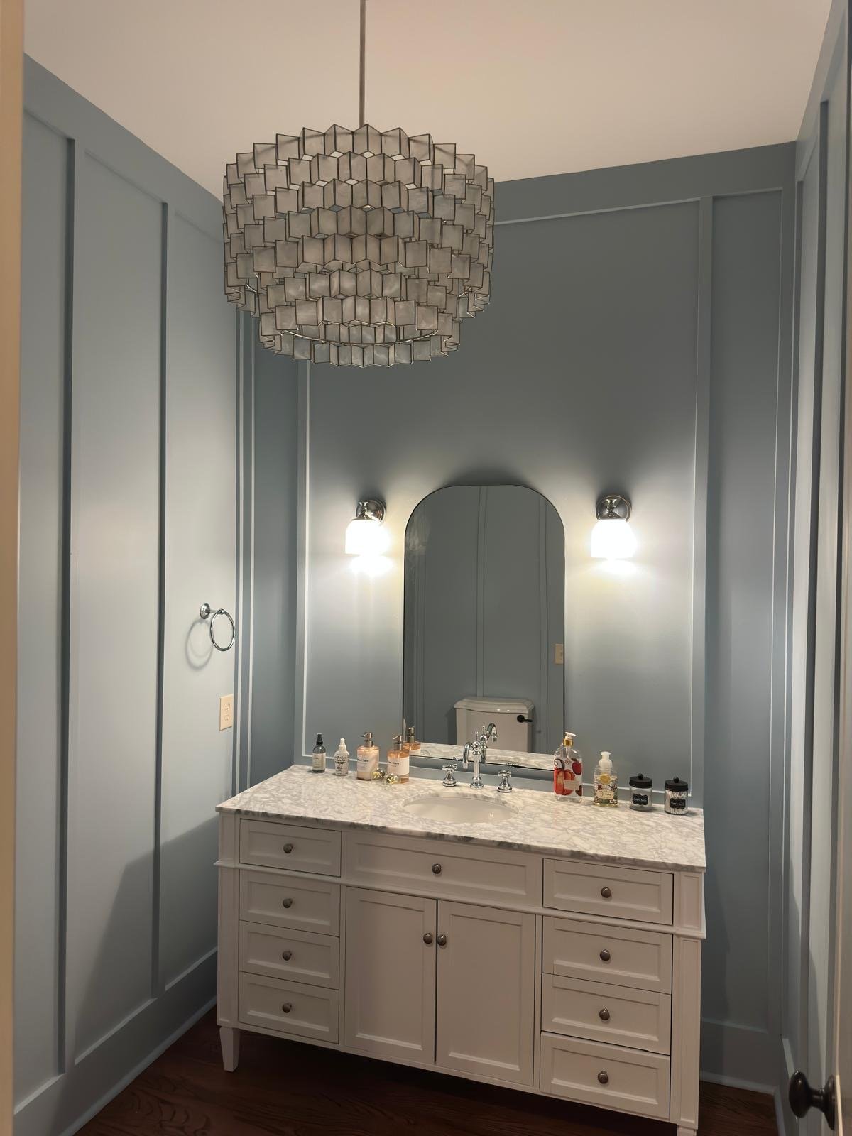 A bathroom vanity with marble countertop, a mirror, and decorative bottles, flanked by two wall-mounted lights, with a patterned ceiling light overhead.