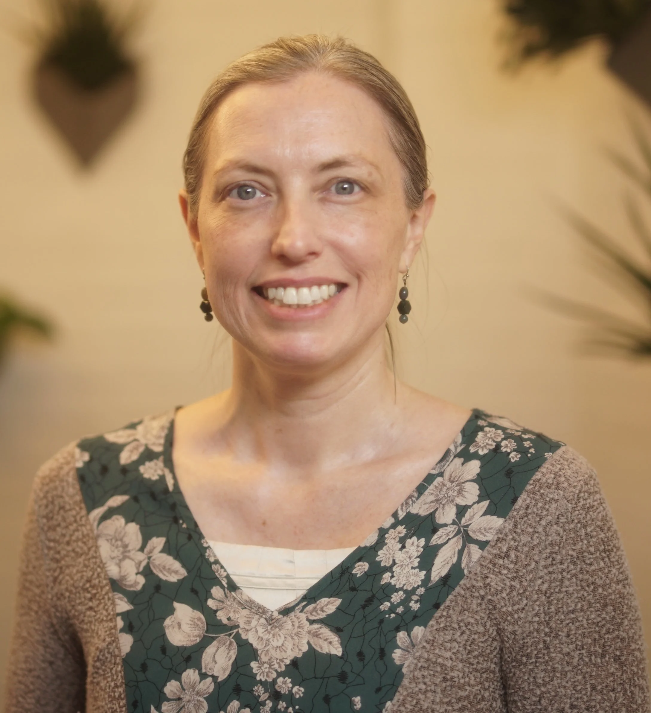 A woman with fair skin, blonde hair, and blue eyes smiling at the camera, wearing earrings and a floral-patterned top. The background is neutral, with some out-of-focus dark plant leaves.