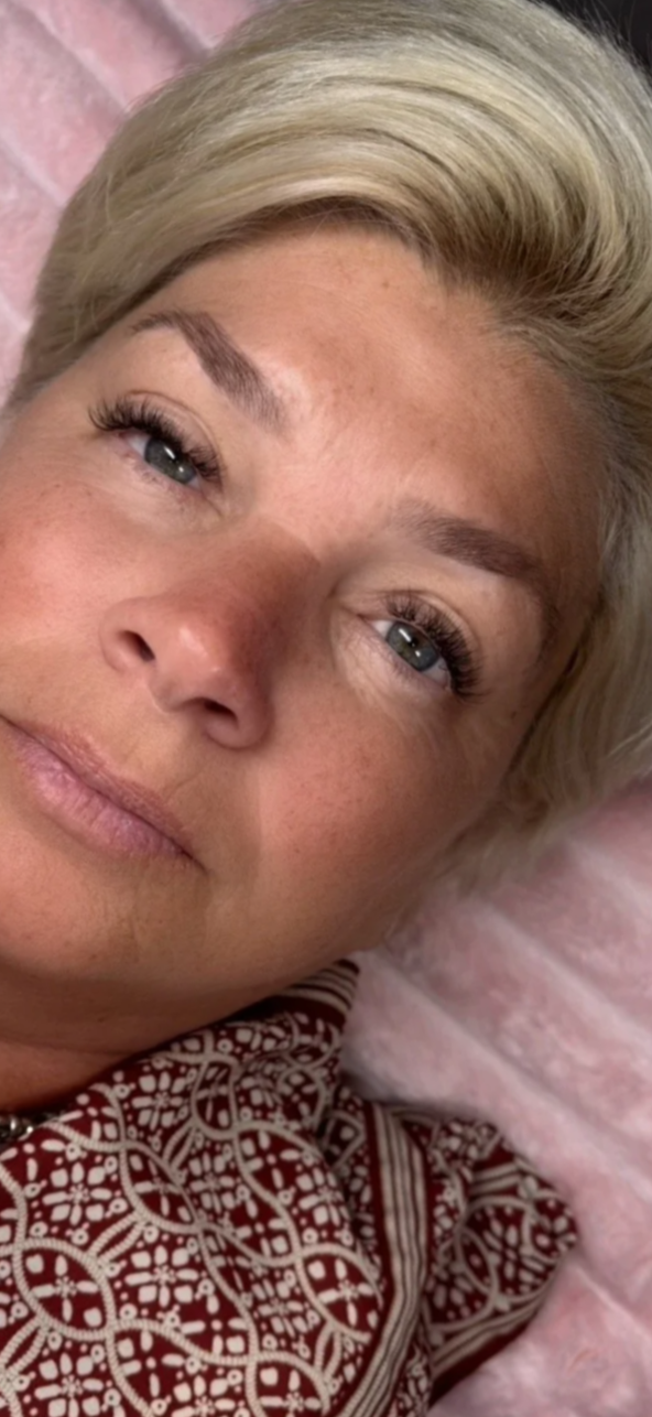 Close-up of a woman's face with blonde hair, blue eyes, and light makeup, lying on a pink fuzzy surface, wearing a patterned top.