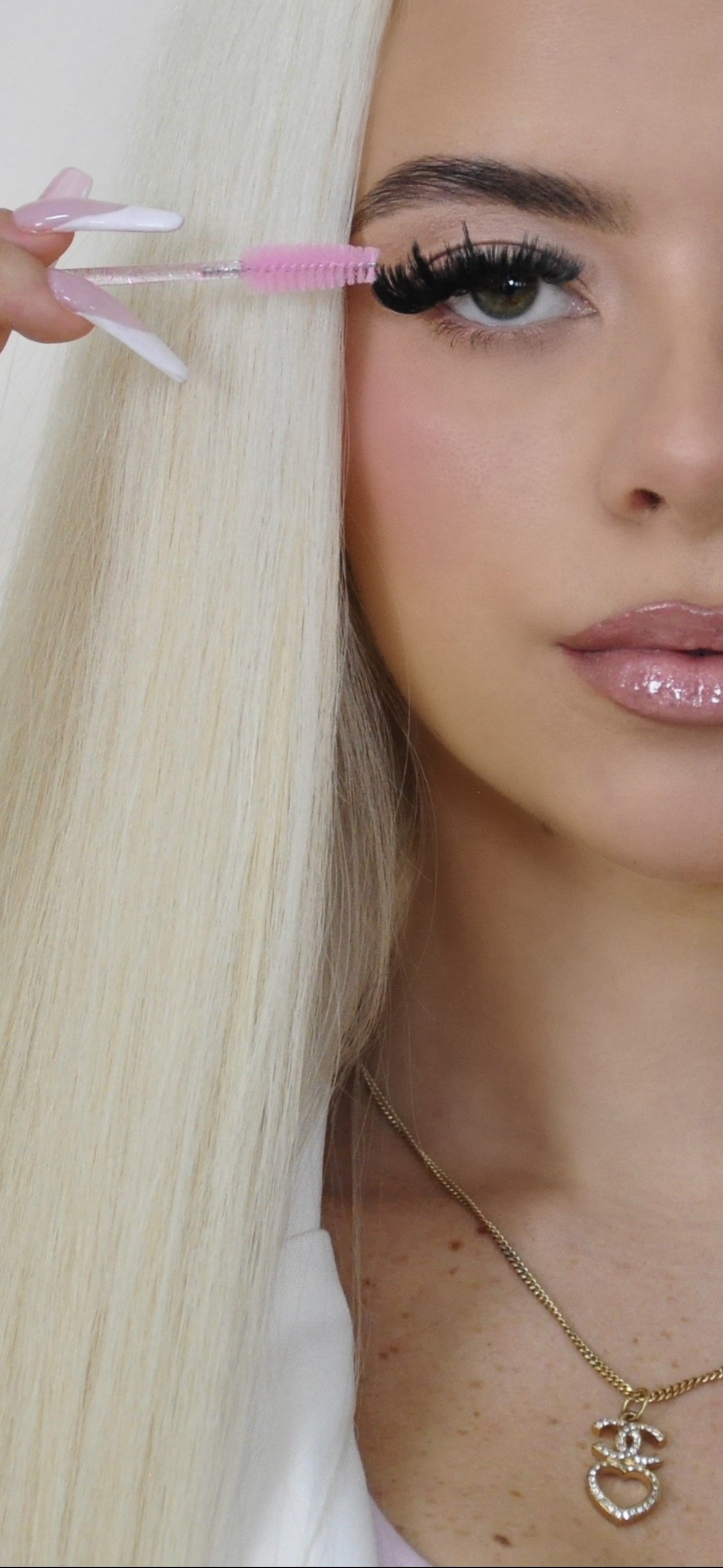 A woman with blonde hair and fair skin is applying pink mascara to her eyelashes with a pink mascara wand. She has a glossy lip with light pink lipstick, subtle eye makeup, and is wearing a gold chain necklace with a decorative pendant. Only part of her face and neck are visible.