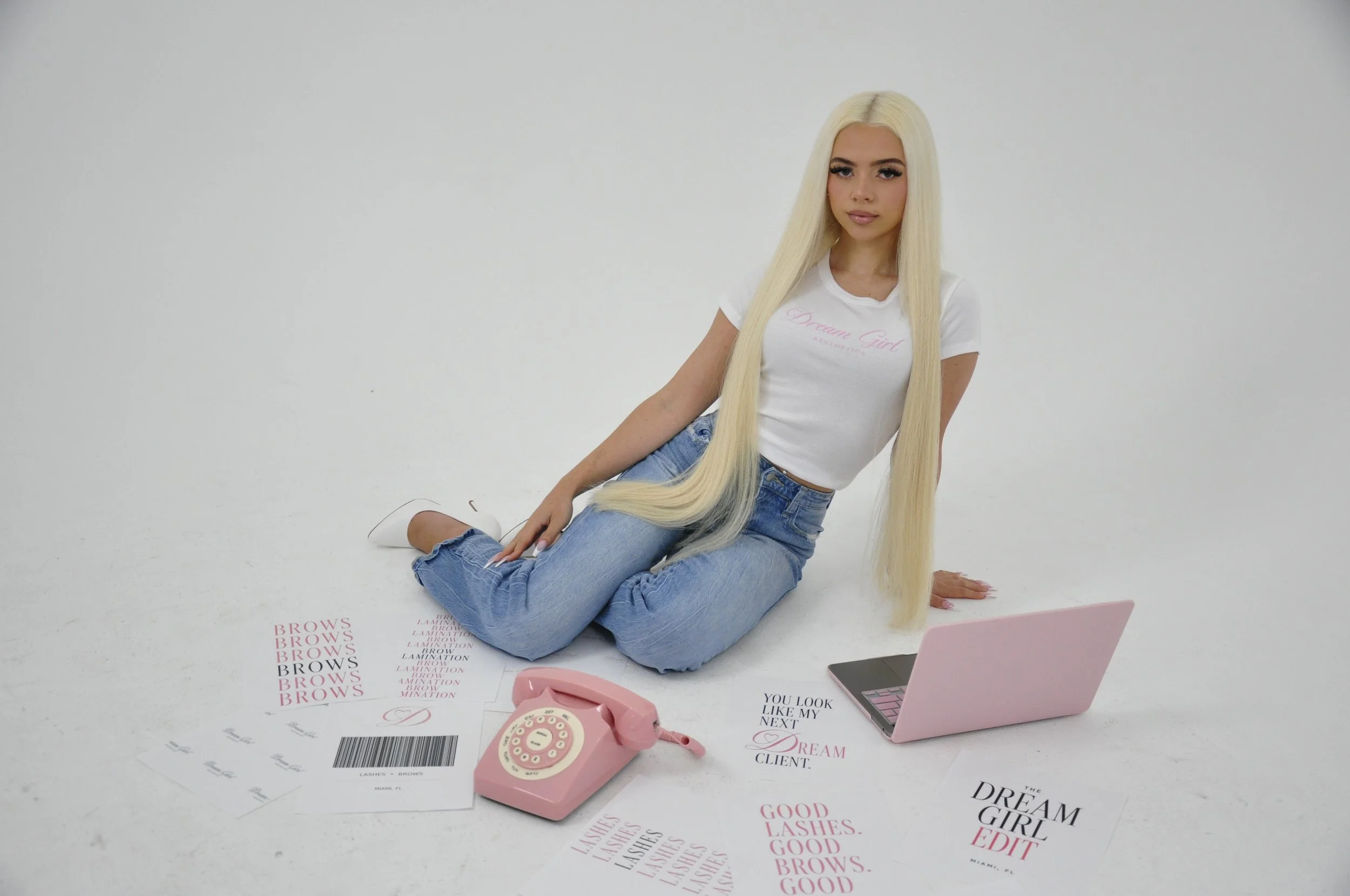 A young woman with long blonde hair and makeup sitting on the floor with a white background. She is wearing a white t-shirt with pink text and blue jeans. There are beauty-related papers, a pink rotary phone, and a pink laptop on the floor around her.