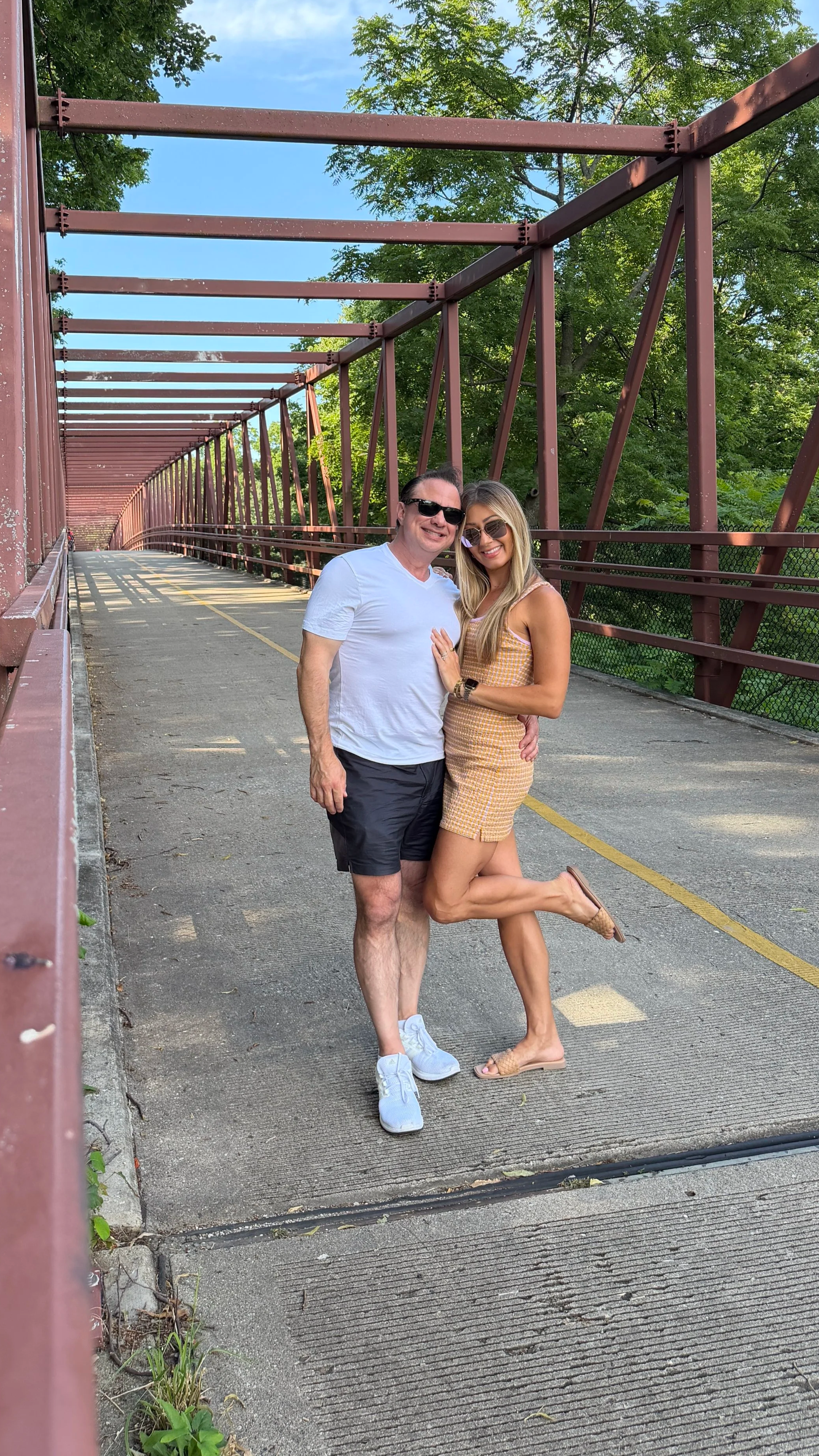 A man and woman standing on a pathway under a red metal bridge, smiling and posing together, with the woman lifting one leg and leaning on the man's shoulder, trees and blue sky in the background.