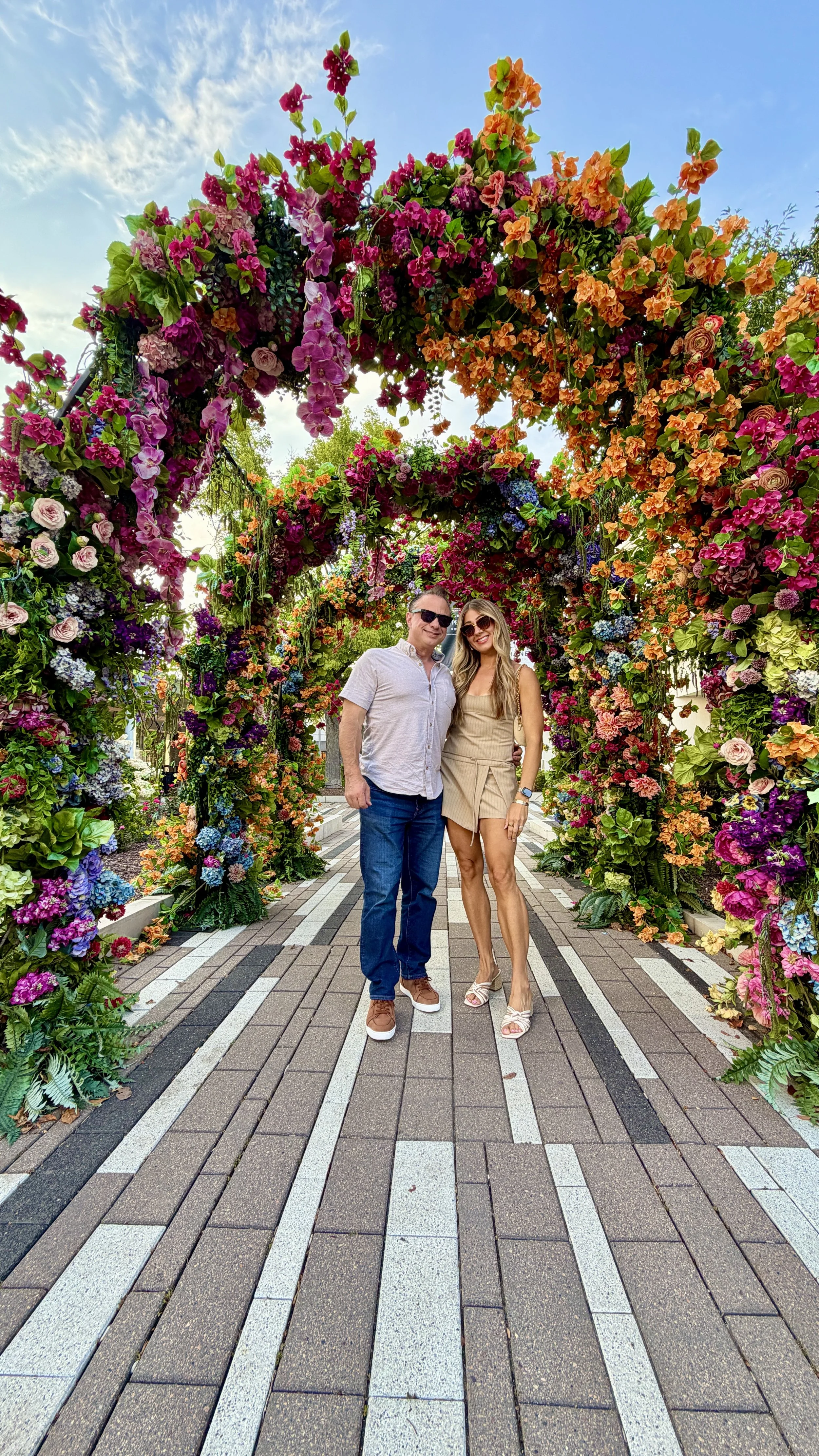 A man and woman standing together under a colorful floral archway in an outdoor setting, with a bright sky overhead.