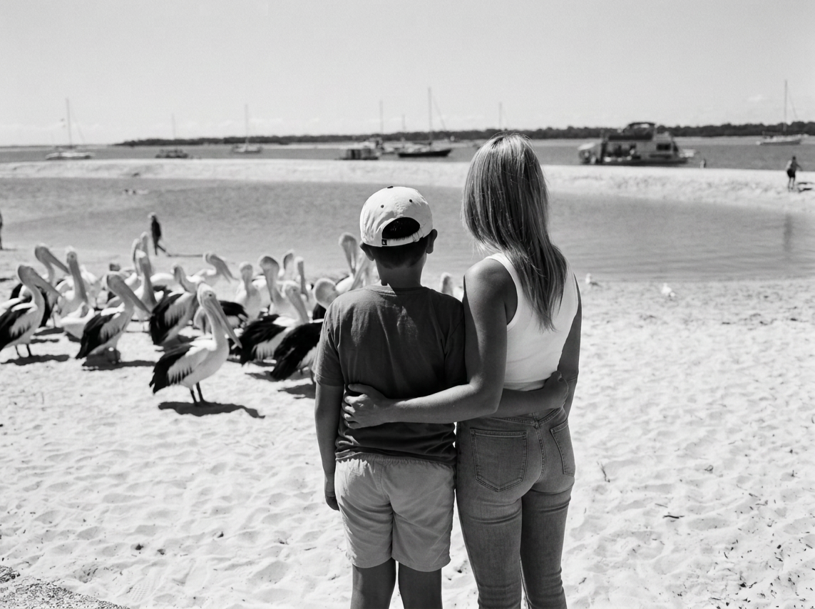 A woman and a boy standing on a sandy beach, facing away from the camera, watching a flock of pelicans with boats in the water in the distance.