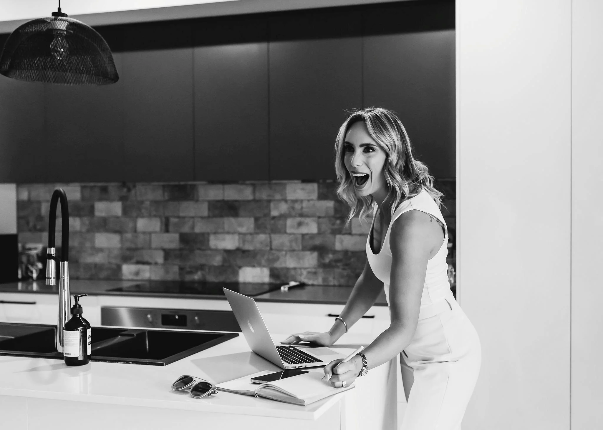 A woman with wavy hair in a sleeveless top working on a laptop in a modern kitchen, smiling and looking excited.