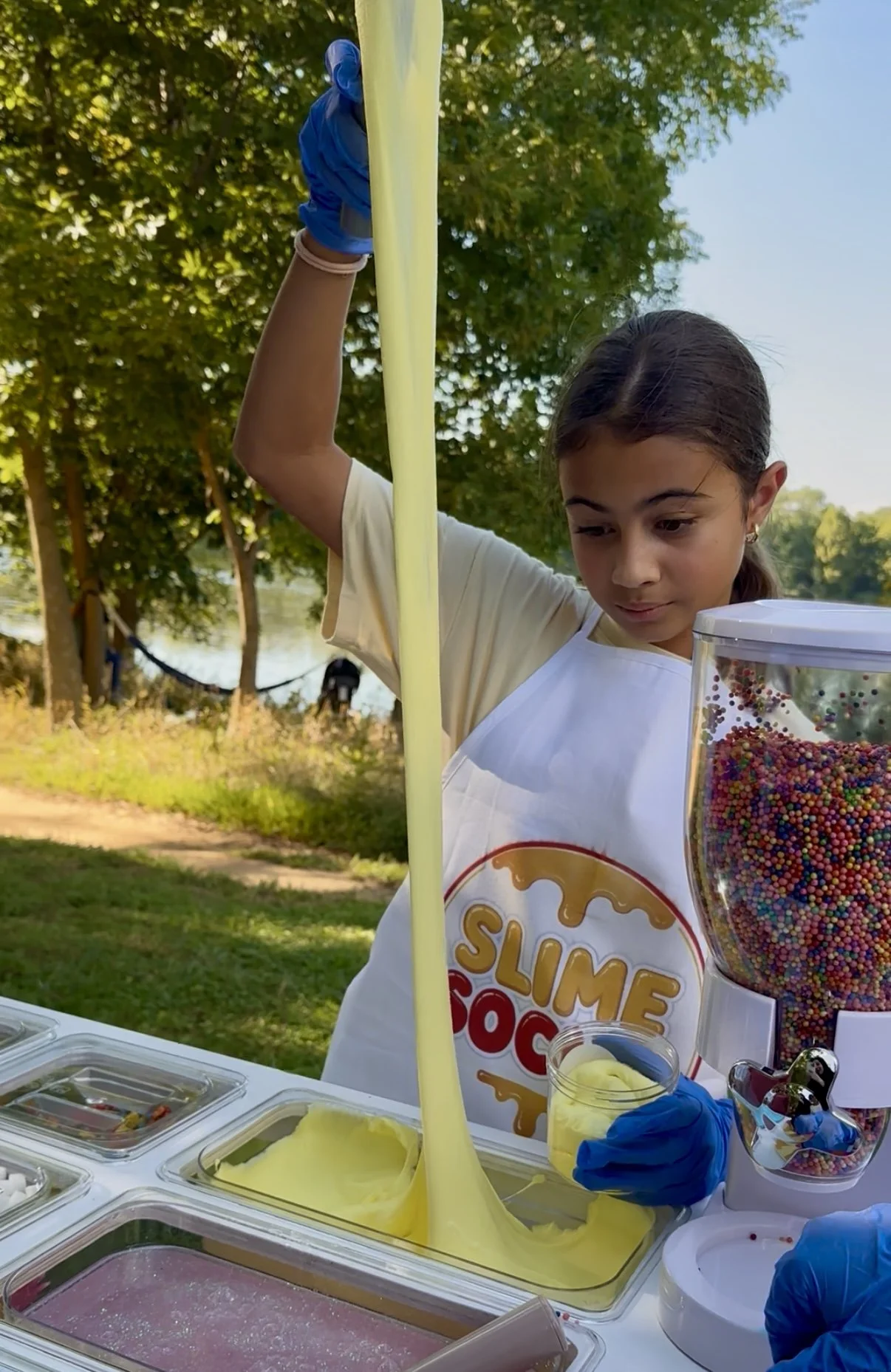 A young girl wearing a white apron and blue gloves at an outdoor slime-making station. She is holding a yellow slime, and a container with colorful sprinkles is visible on the table. The background features green trees and a lake or river.