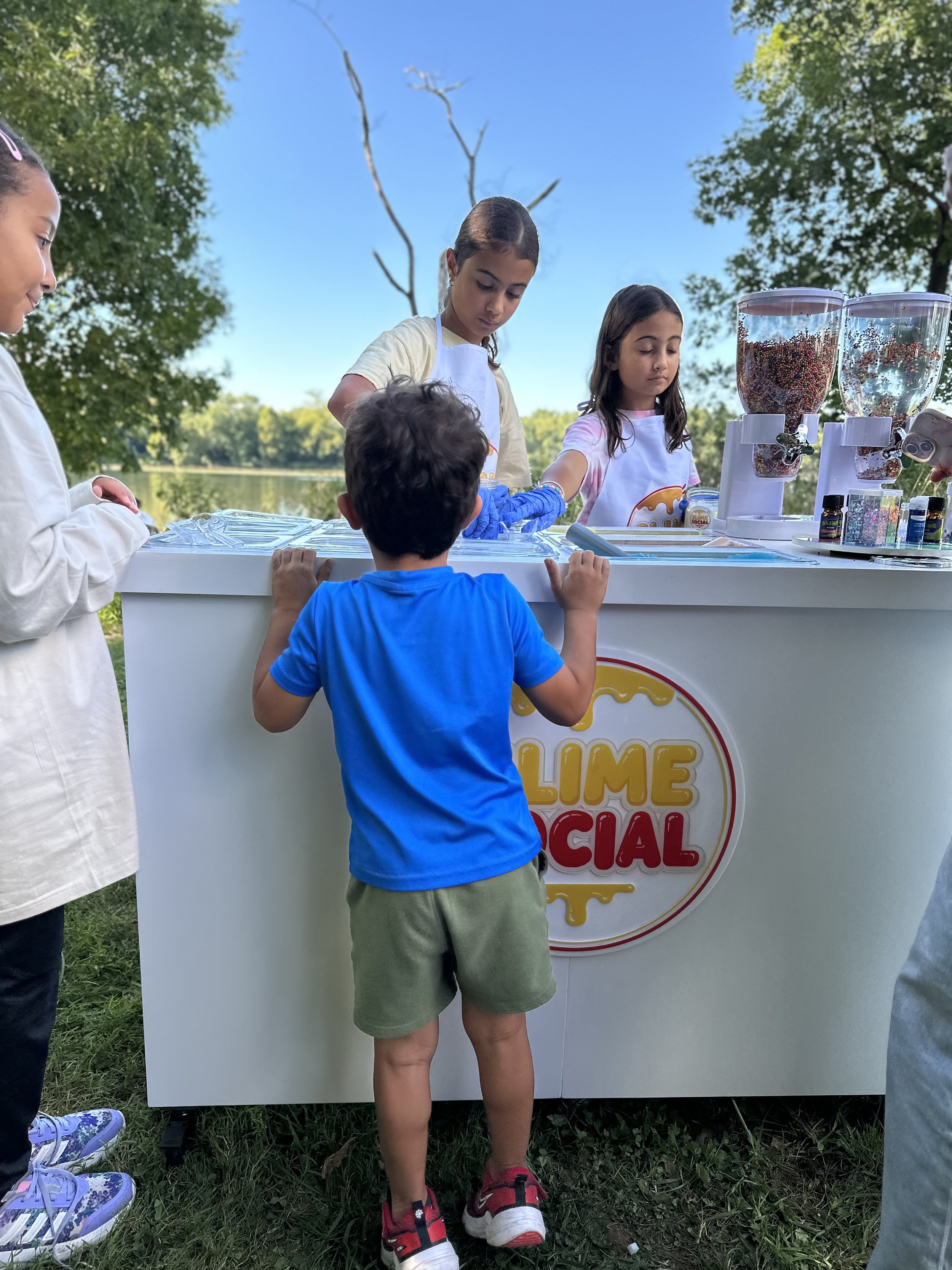 Children at a lemon-themed ice cream stand outdoors near a lake, wearing aprons, with two large containers of colorful sprinkles on the counter.