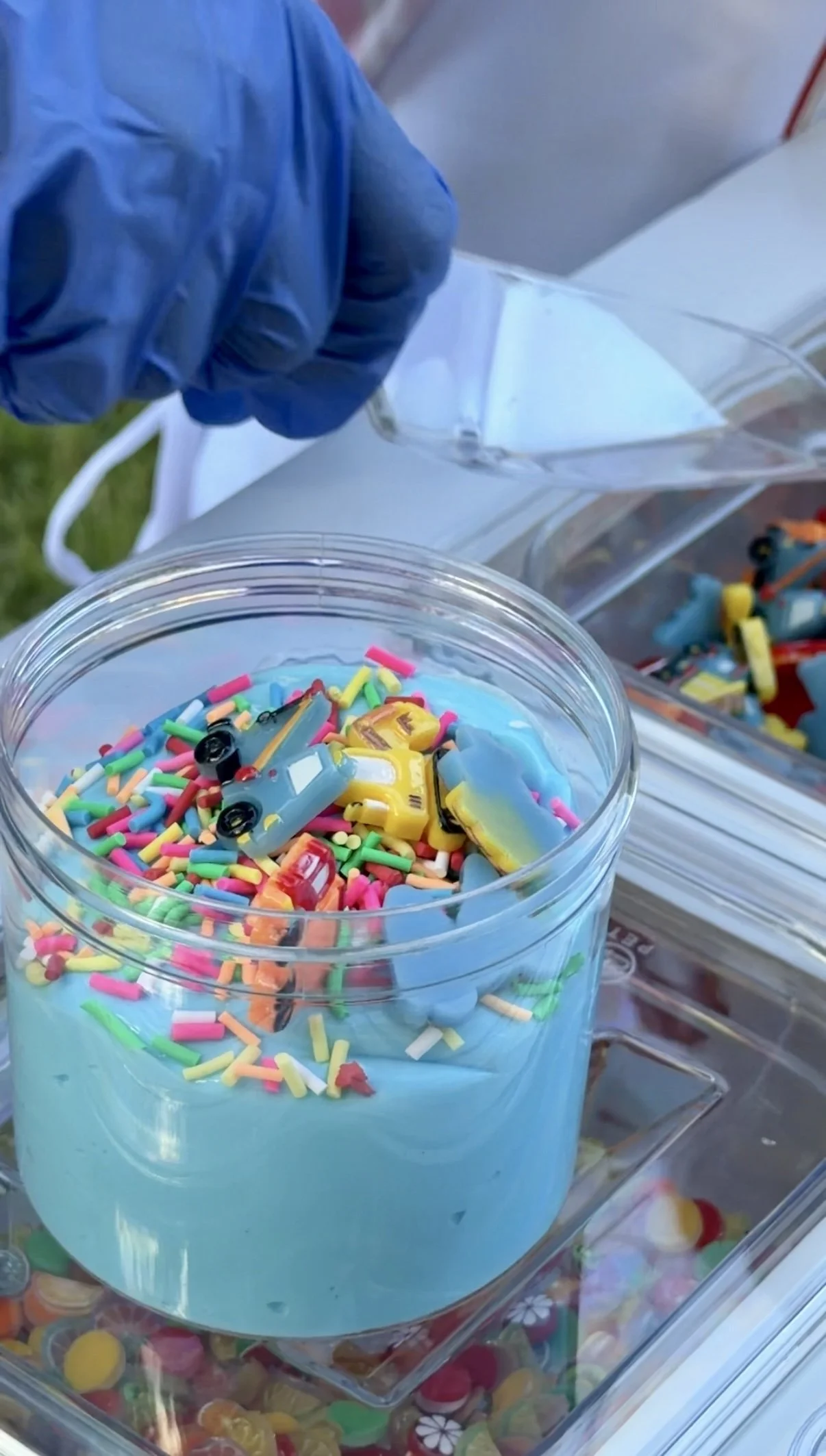 Container of colorful sprinkles and toy cars on display at a candy store.